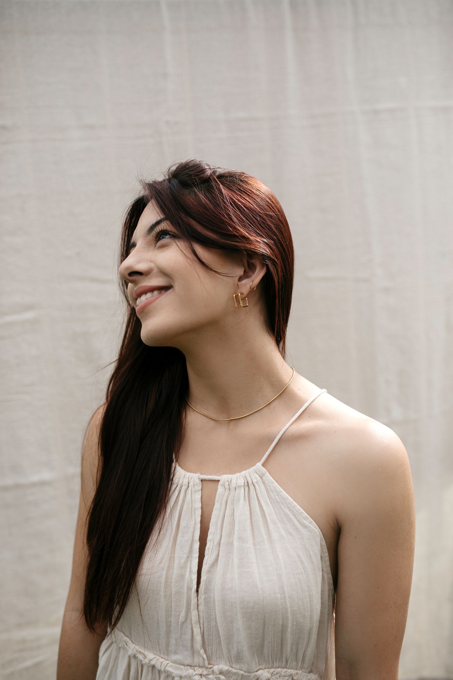 A person in a white halter dress smiles while looking up, wearing a delicate necklace against a light background.