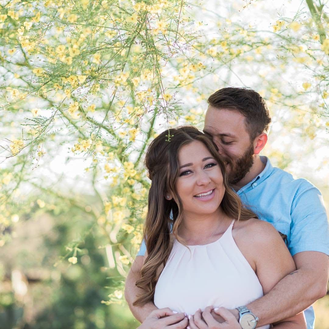 A romantic couple embraces under blooming yellow trees in soft natural lighting during an outdoor photoshoot.
