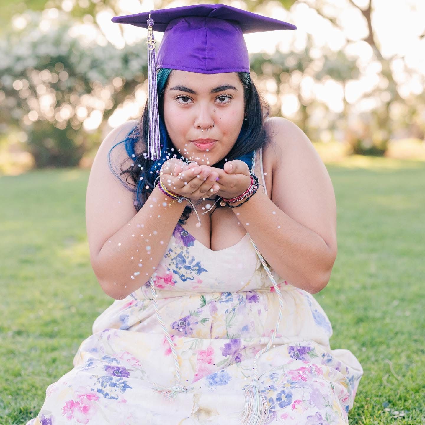 Graduate in purple cap blowing glitter in a floral dress outdoors.