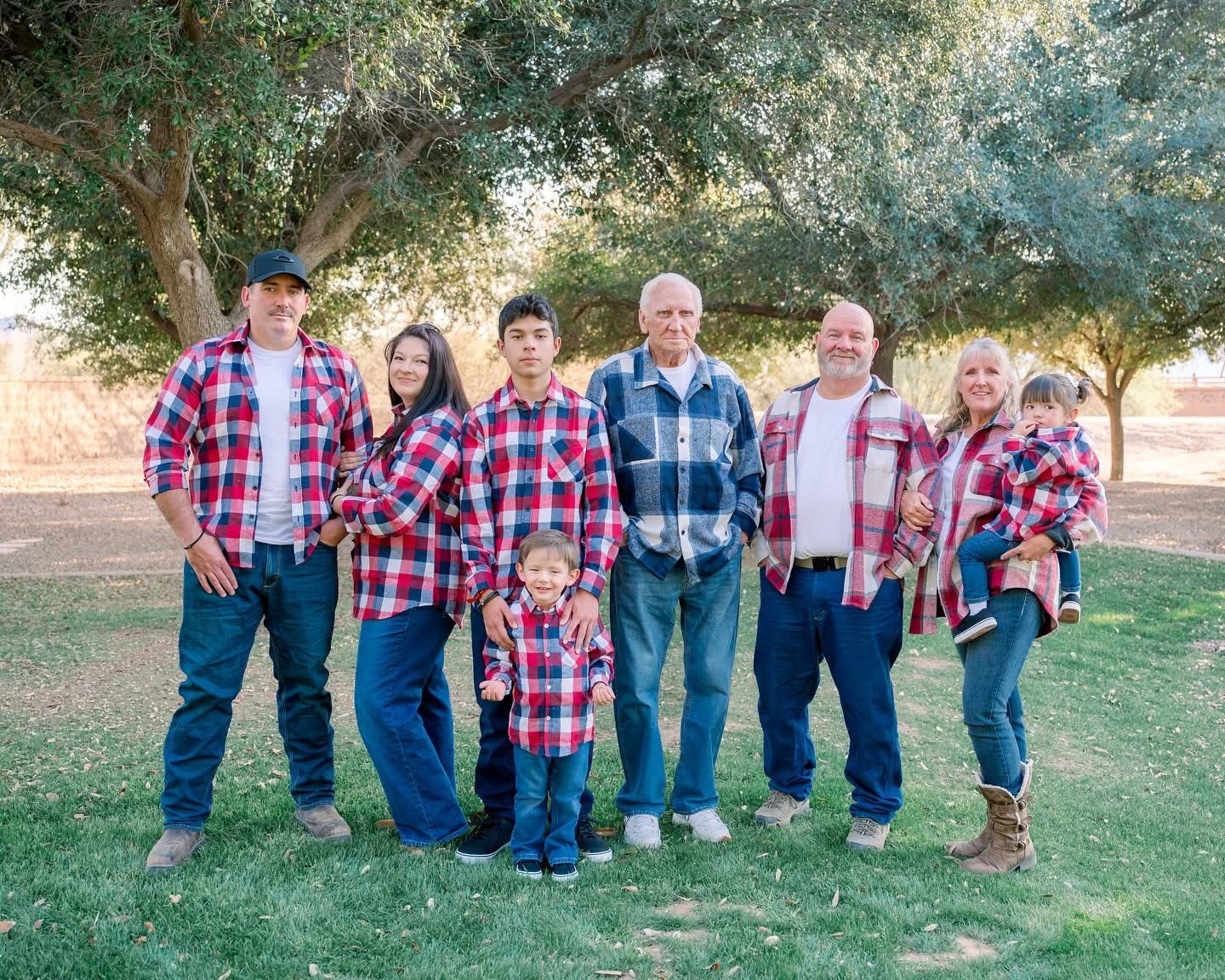 A family group wearing coordinating red and blue plaid shirts poses together outdoors under trees in a park setting.