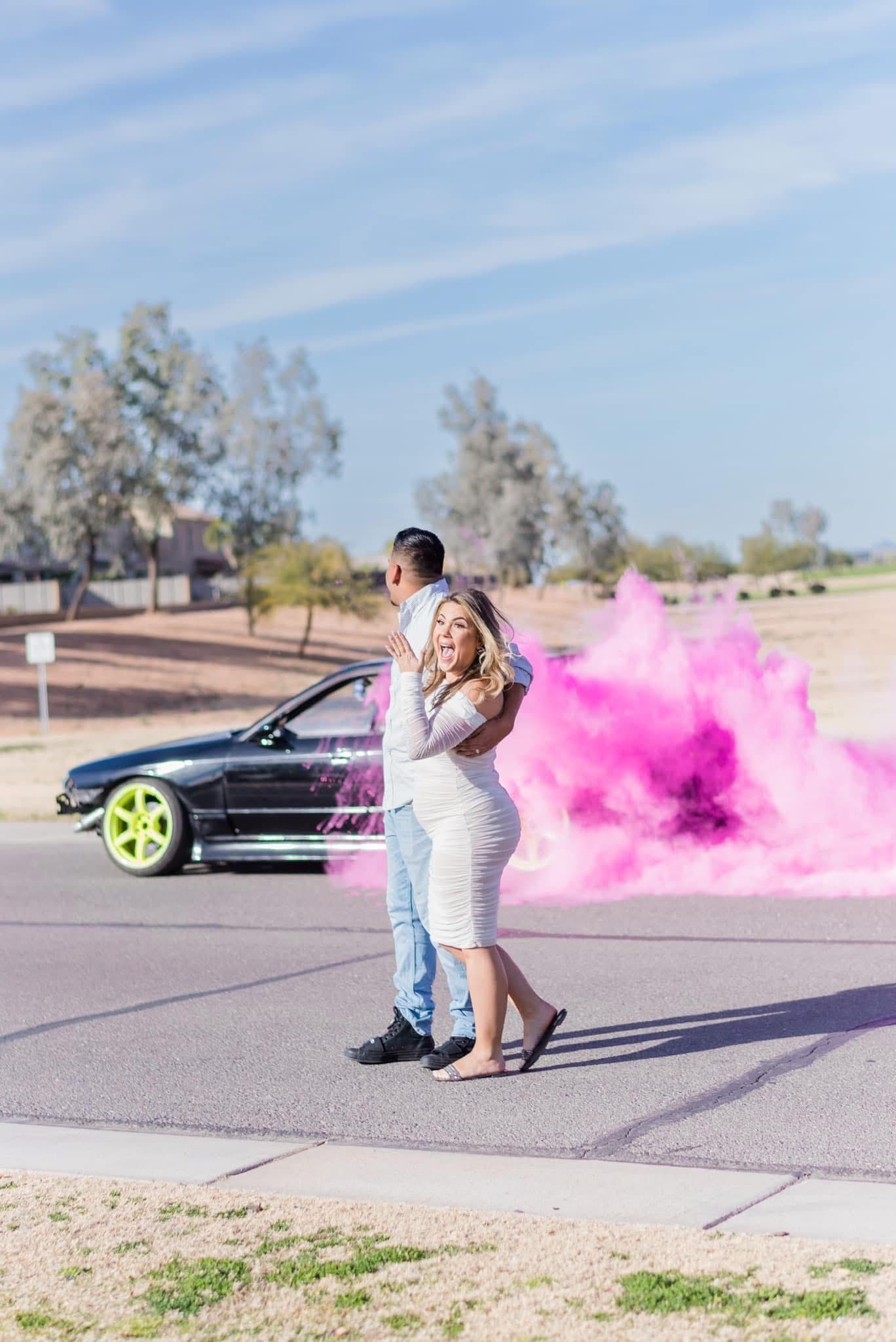 A couple poses together with pink smoke and a black car in the background on a desert road.