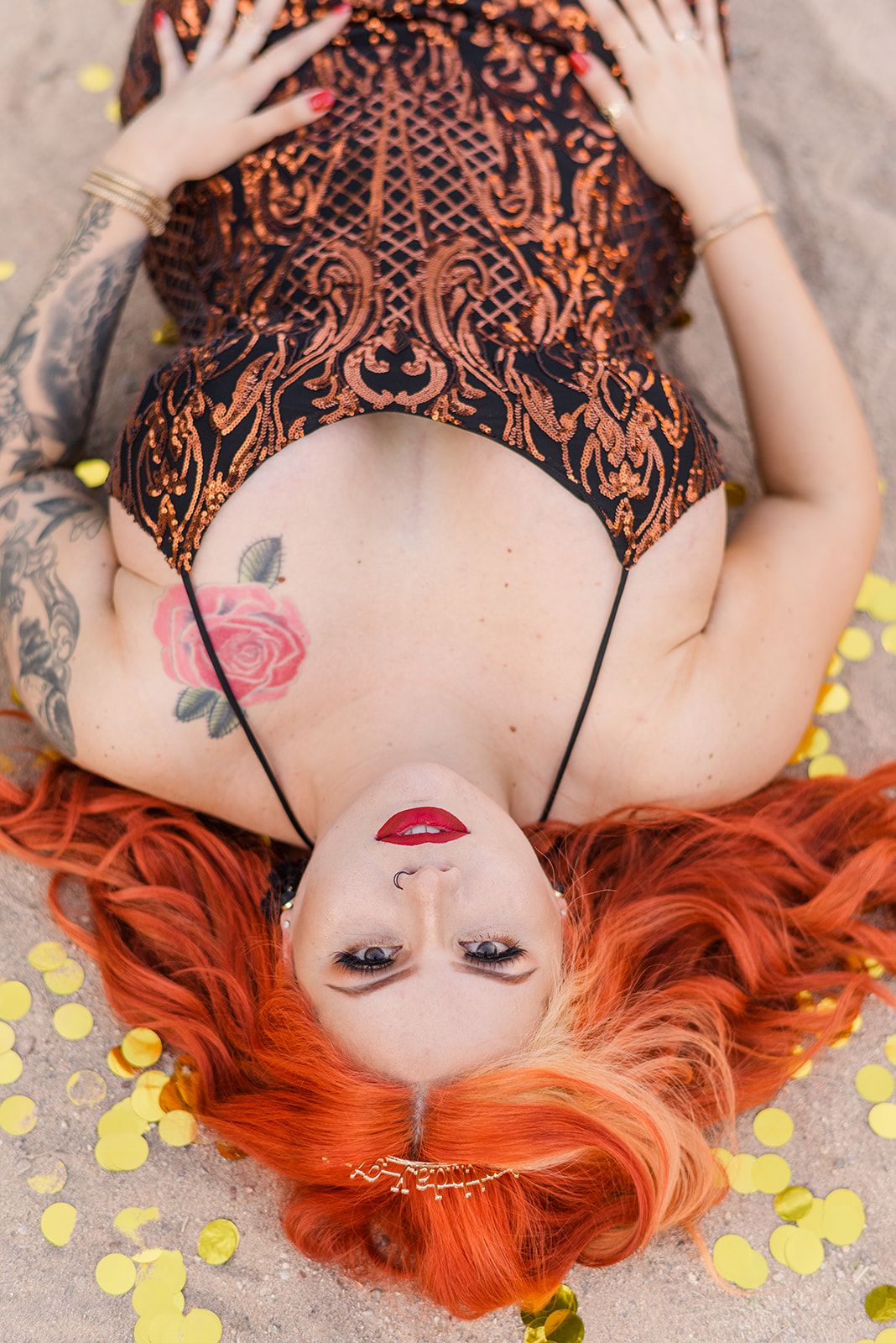 Portrait of person with bright red hair lying on sand surrounded by yellow petals wearing ornate clothing.
