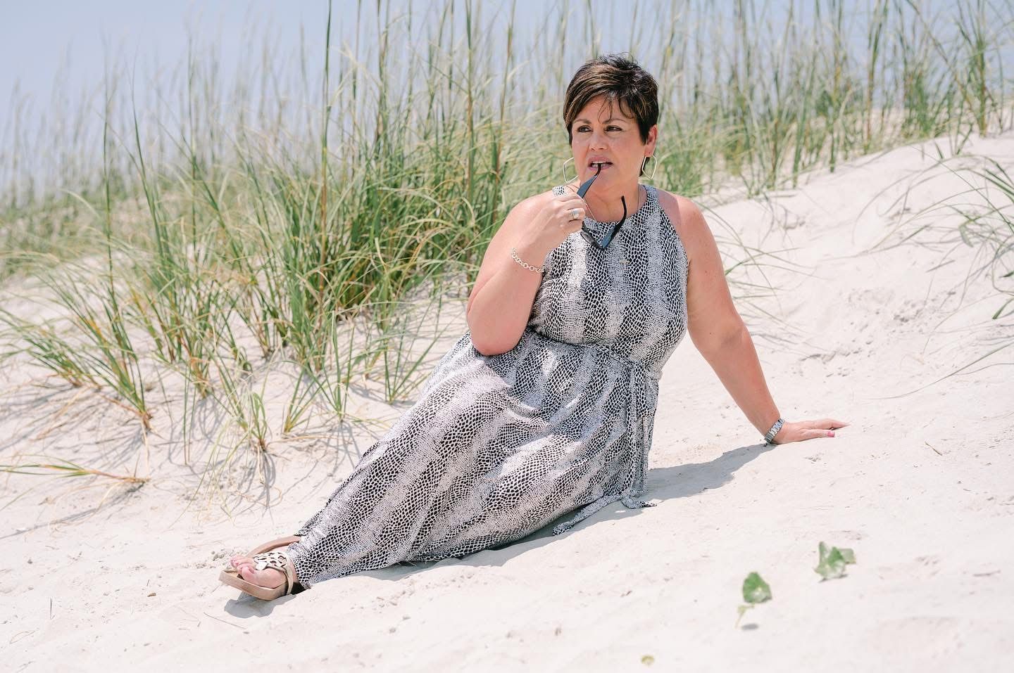 Woman in flowing gray dress sits gracefully on sandy beach dunes.