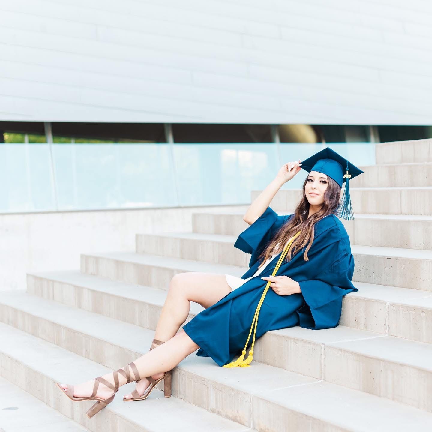 Graduate in blue cap and gown sitting on concrete steps for graduation photos.