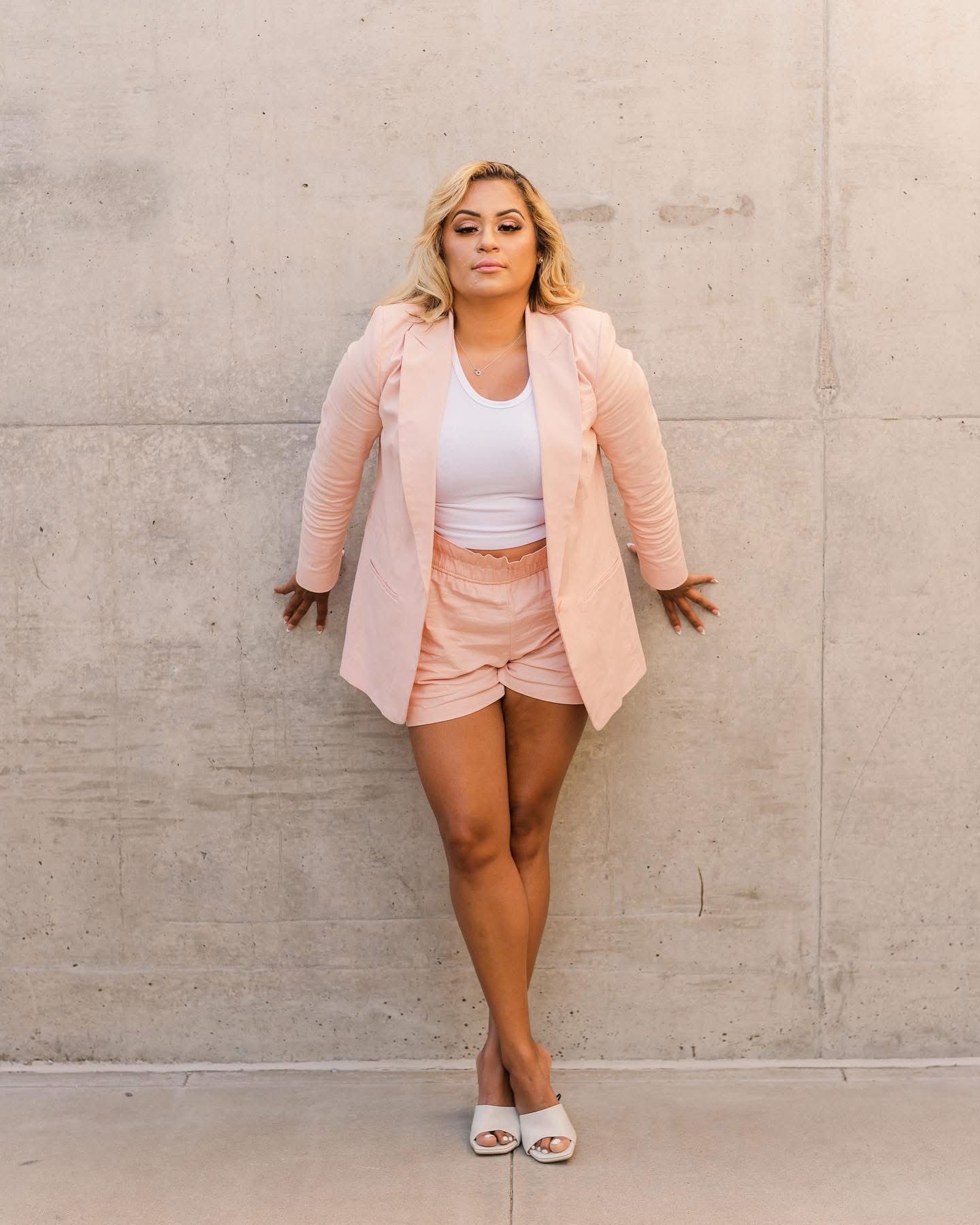 Fashionable woman in pink blazer and shorts poses confidently against concrete wall.