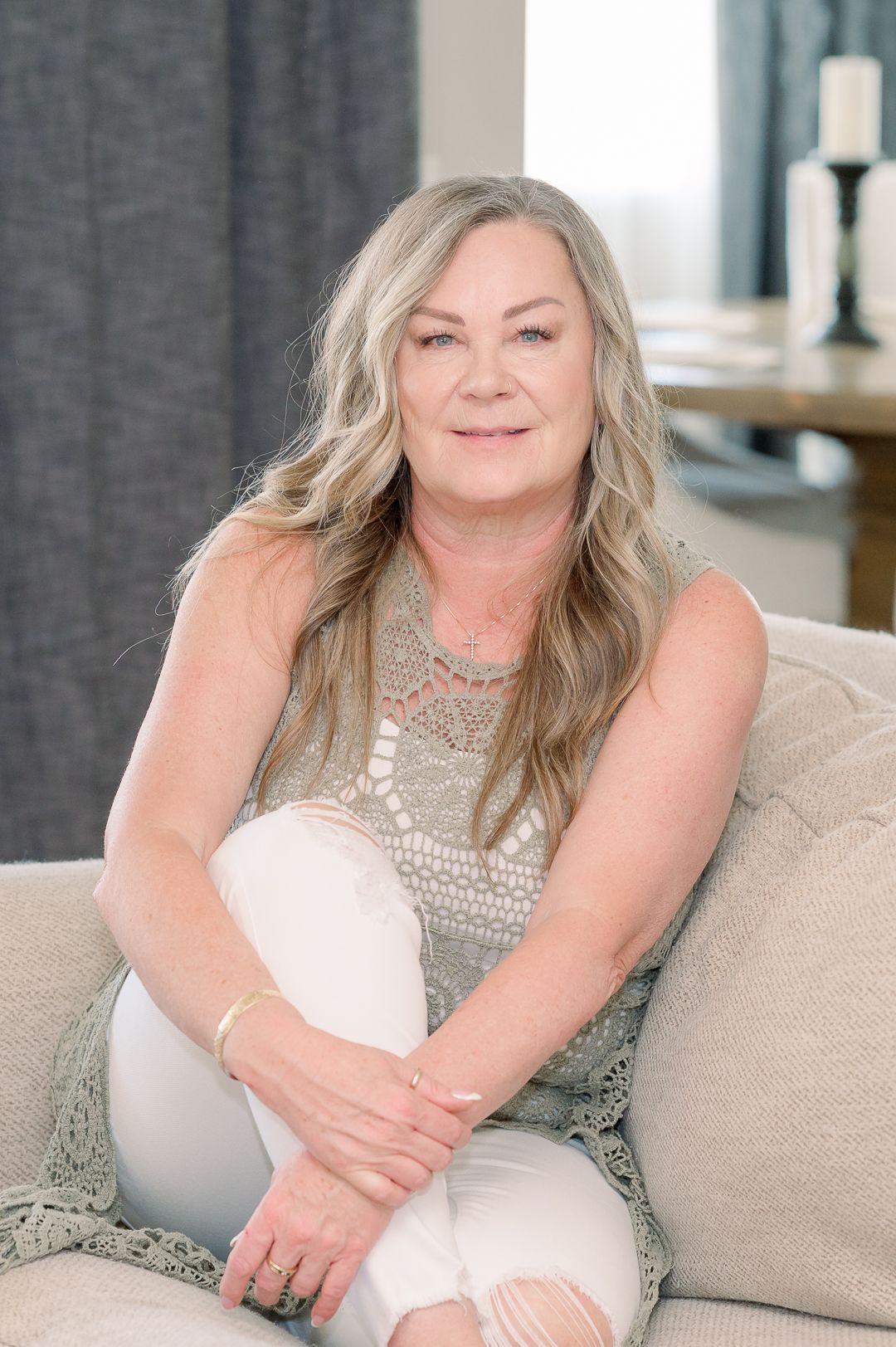 A person in a silver lace top sits comfortably on a beige couch with long blonde wavy hair and relaxed posture.