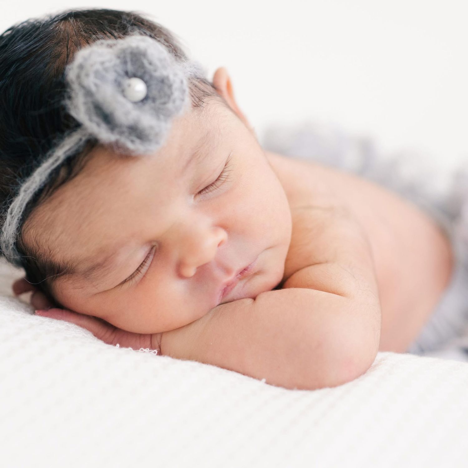 A sleeping newborn baby wearing a grey flower headband rests peacefully on white bedding.