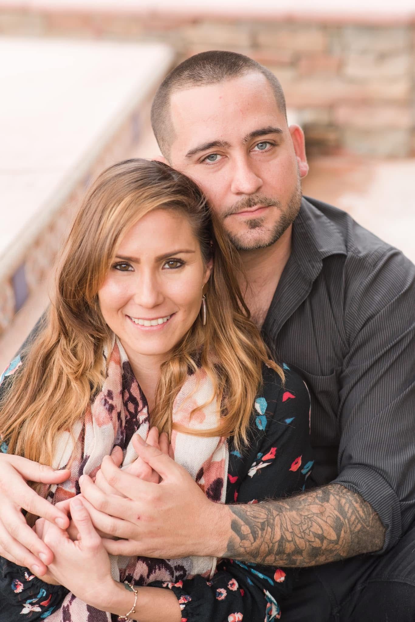 A couple in casual attire poses intimately for a romantic portrait against a brick wall.