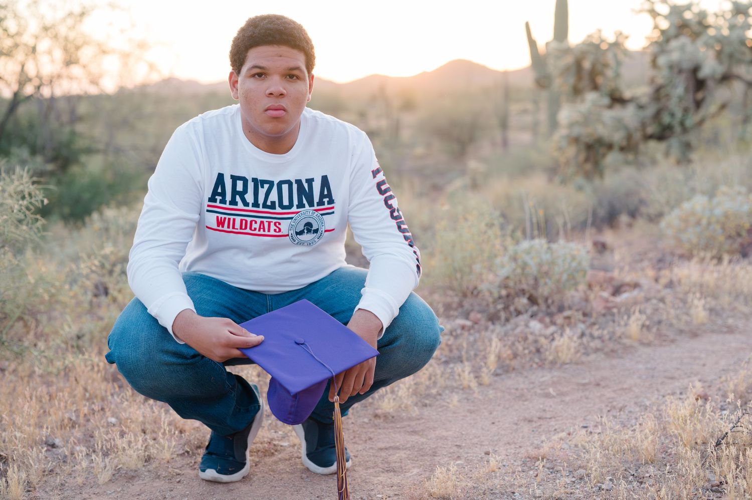 Person in an Arizona sweater crouching in a desert landscape at sunset.