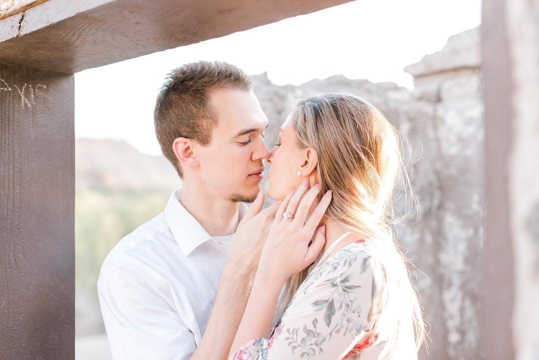 A romantic couple shares a tender kiss in soft sunlight against an architectural backdrop.
