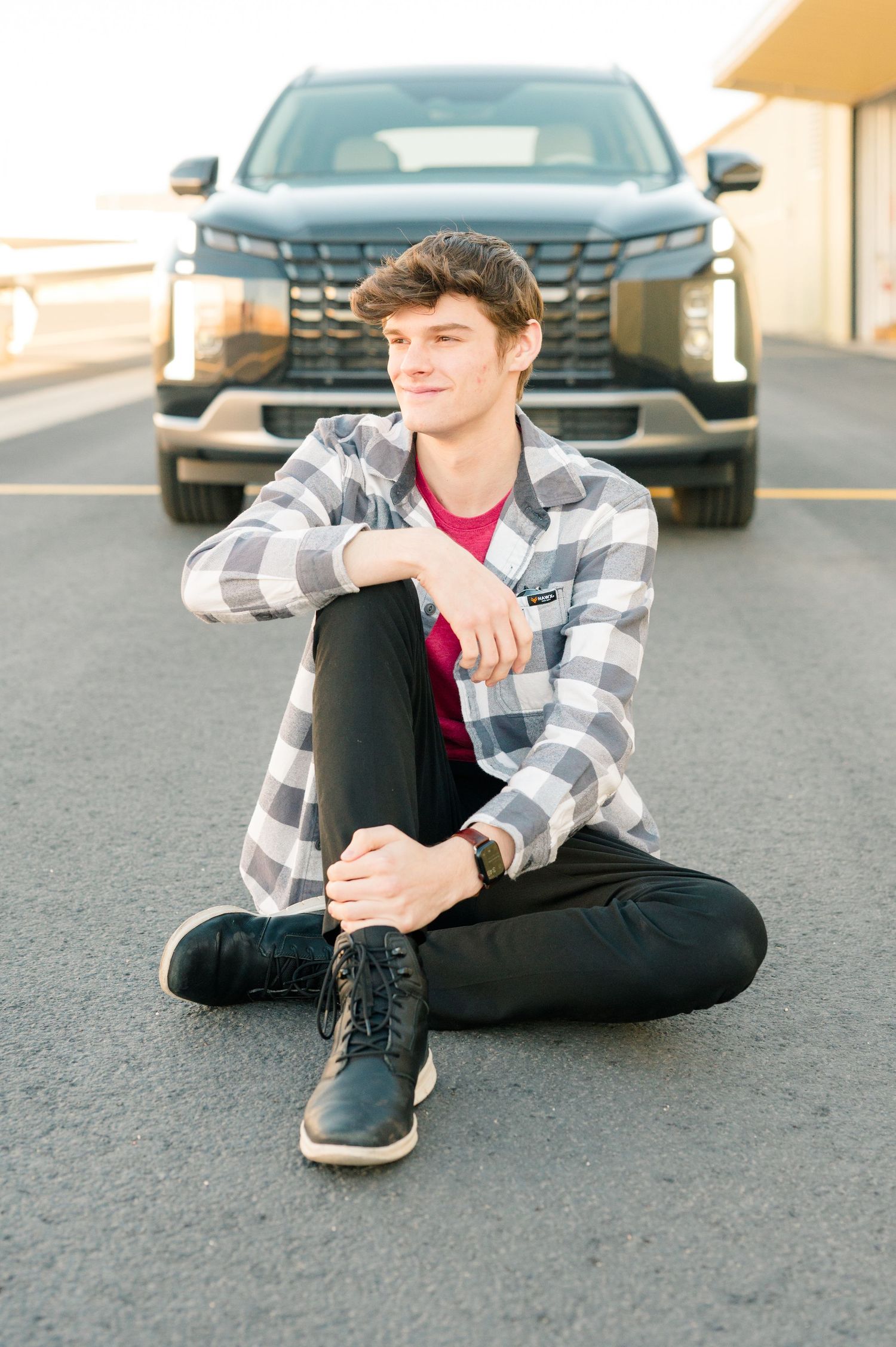 Person sitting on pavement wearing plaid shirt and black pants in parking lot.