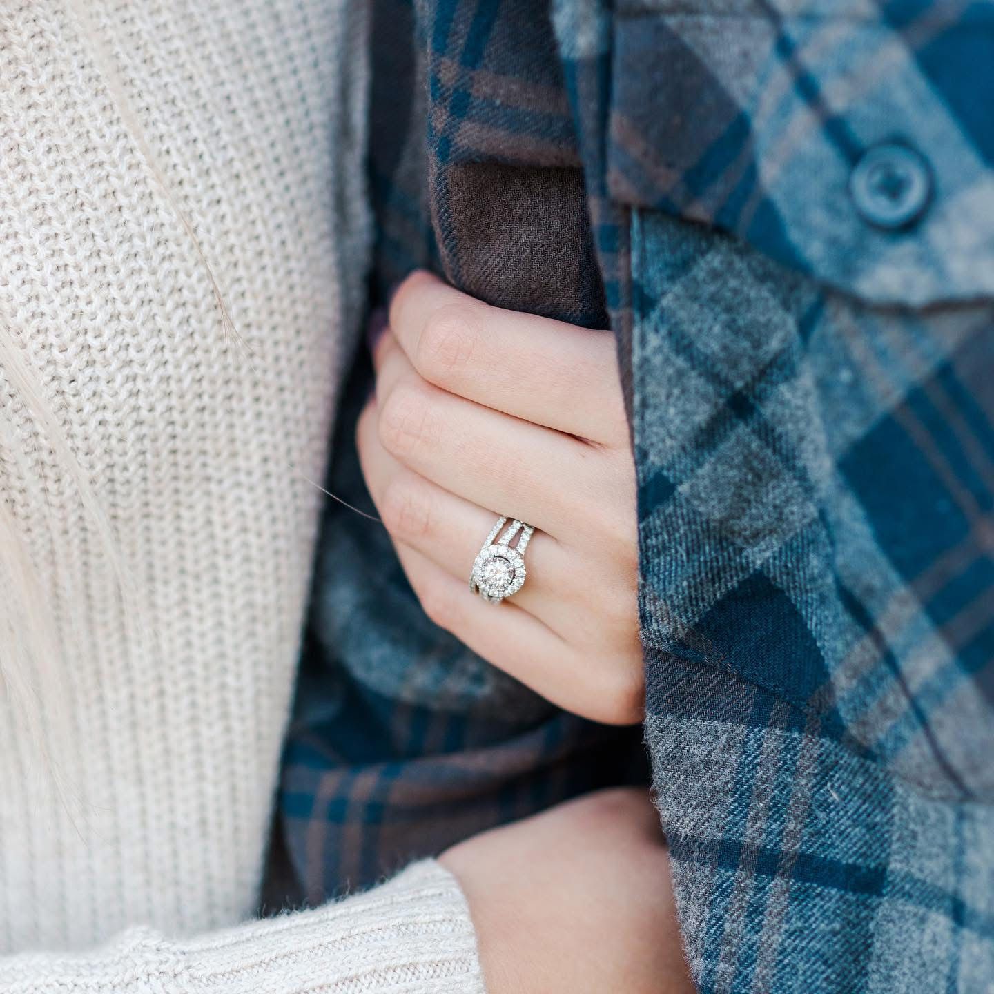 Close up of a diamond engagement ring worn with a white sweater and plaid shirt.