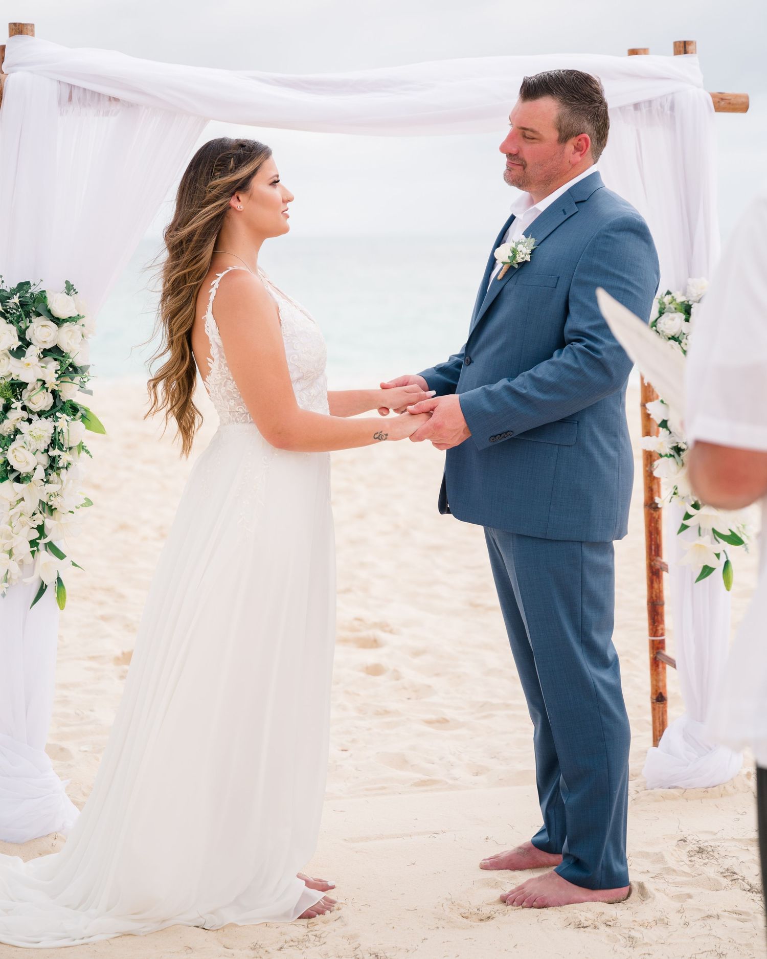 Beach wedding ceremony under white arch with floral arrangements on sandy shore.