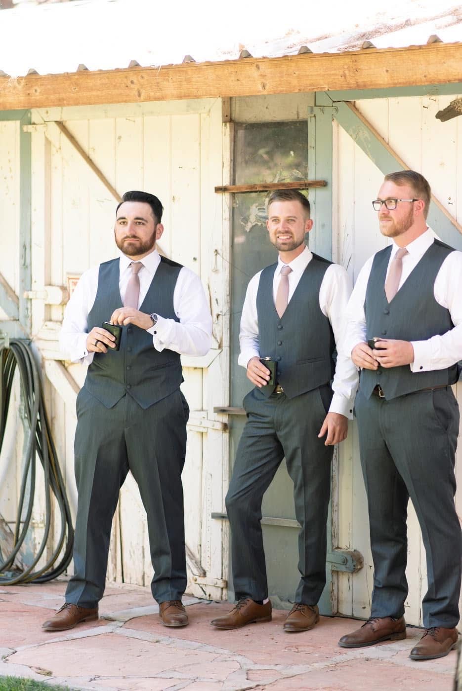 Groomsmen in matching gray vests and brown shoes lean casually against rustic wall during outdoor wedding photo shoot.