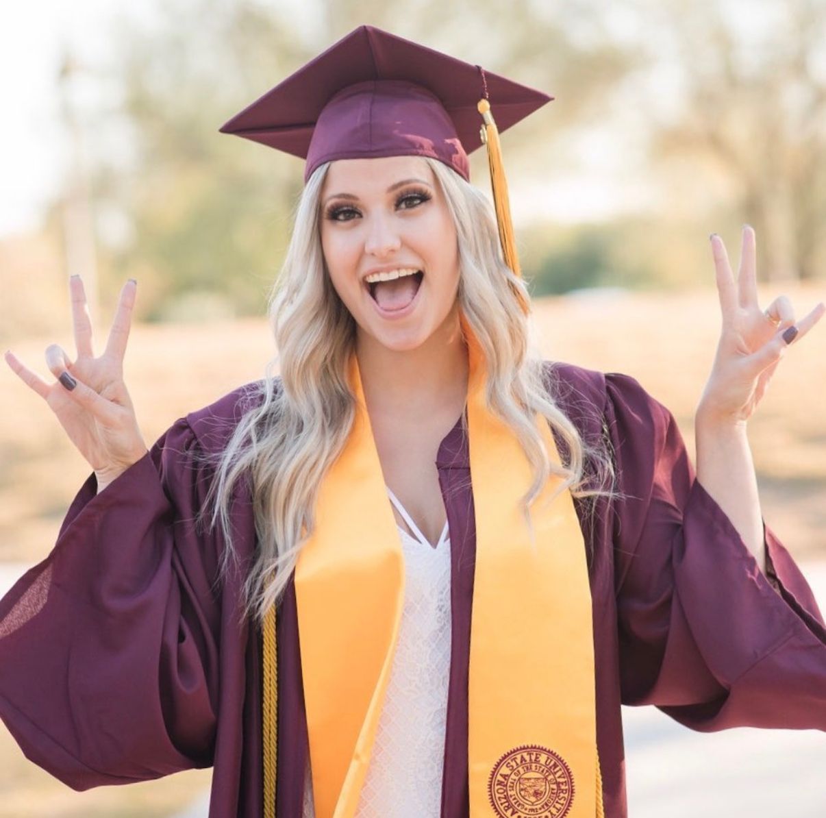 Graduate in maroon cap and gown with yellow stole showing peace signs with excited expression.