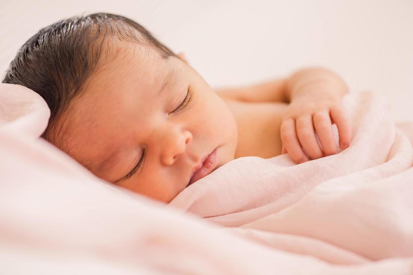 Peaceful newborn baby sleeping under soft pink blanket in gentle natural light.