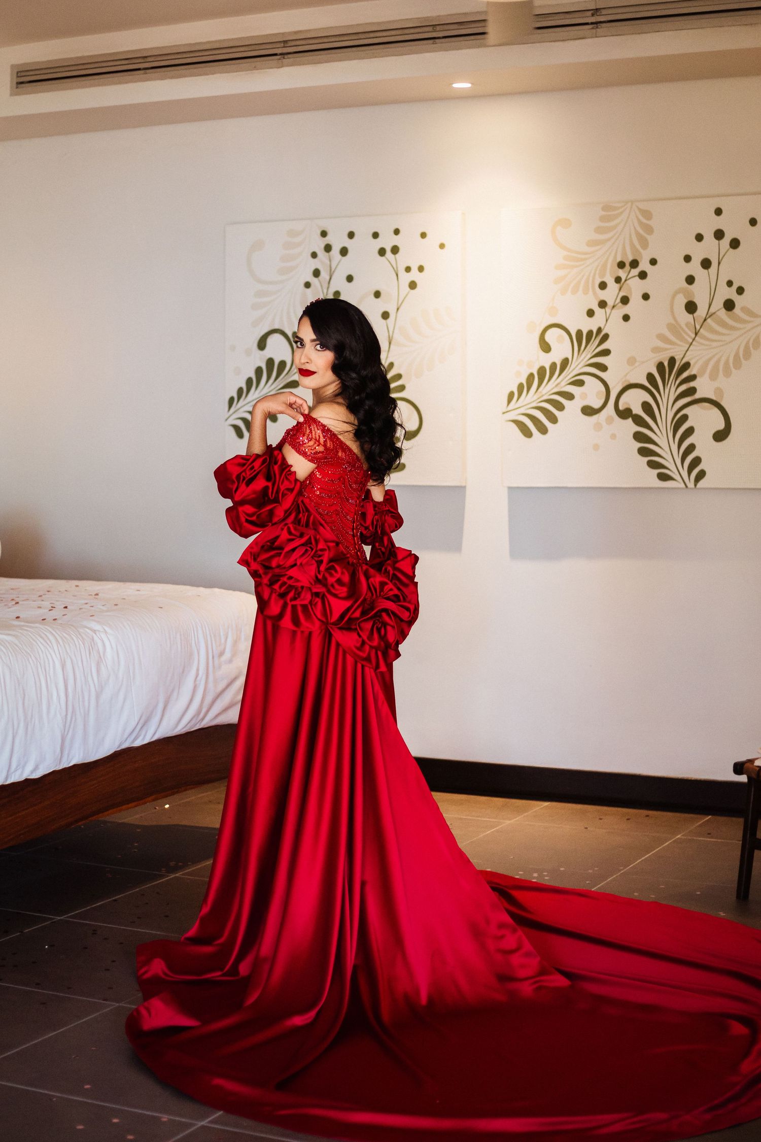 A woman in a dramatic red ball gown with ruffled details poses in an elegant room with floral wall decorations.
