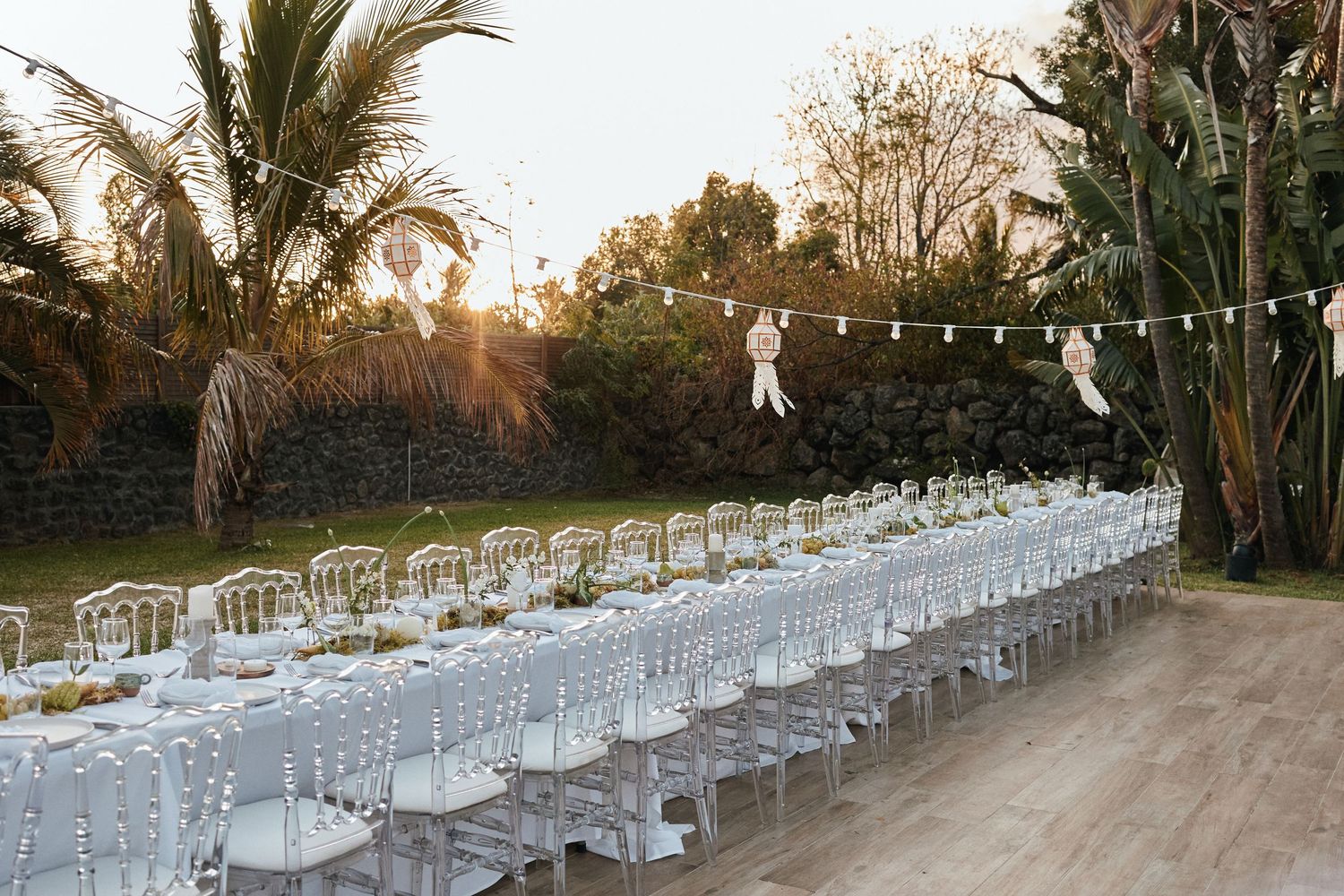 Elegant outdoor wedding reception setup with long table and clear chairs under string lights.