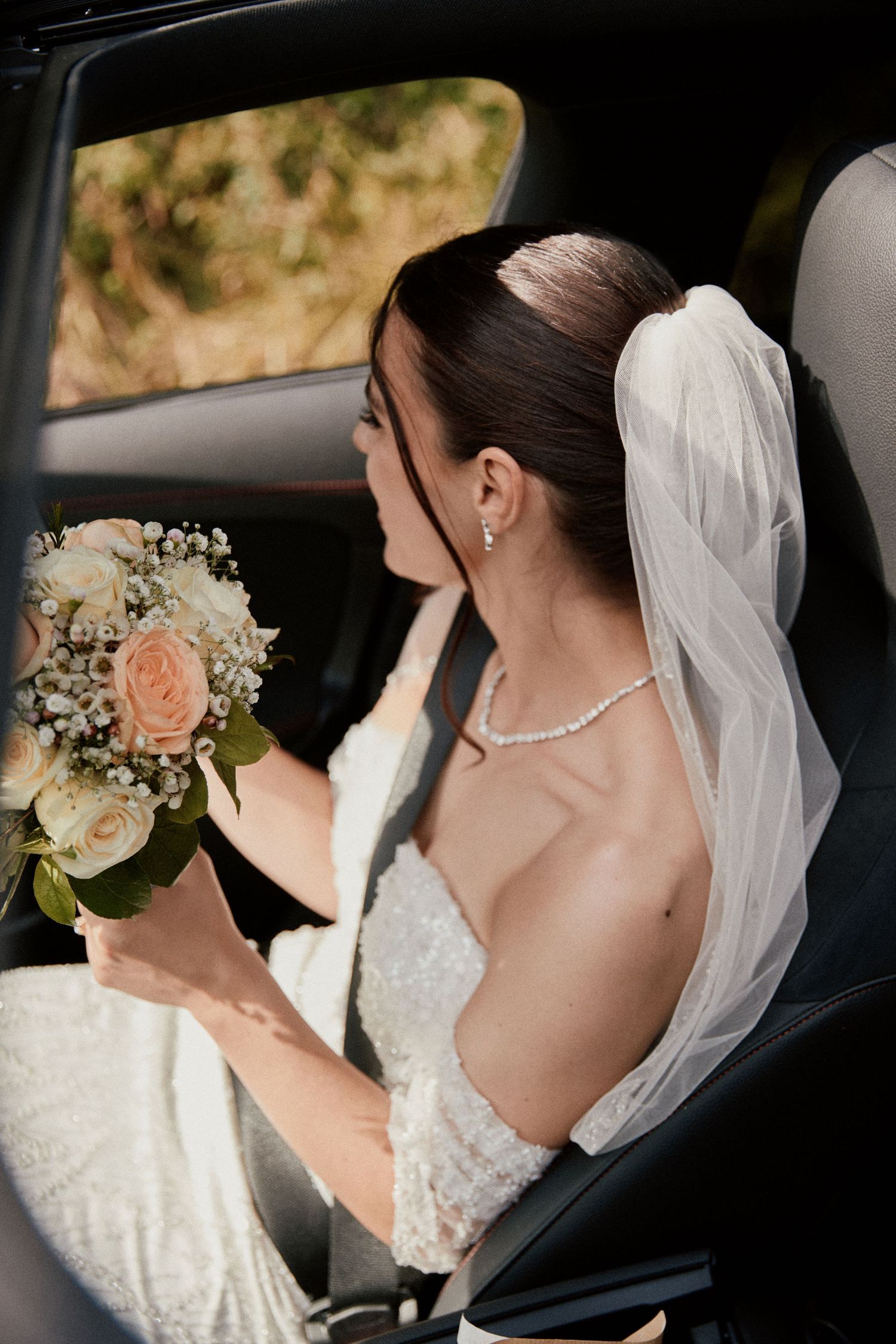 A bride sitting in a car holding a peach and white bouquet with her veil flowing behind.