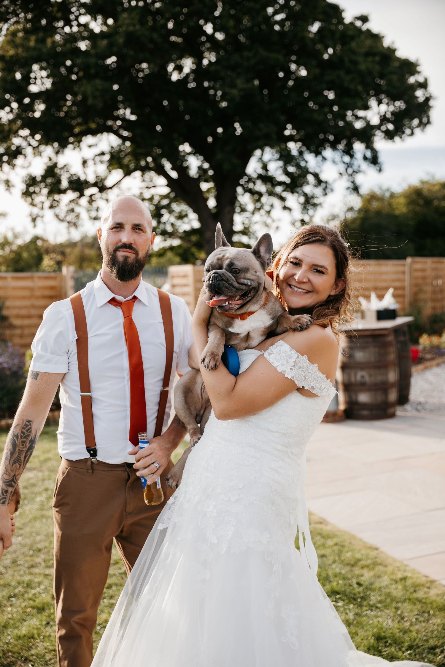 Newlyweds in casual wedding attire pose with their French Bulldog outdoors.