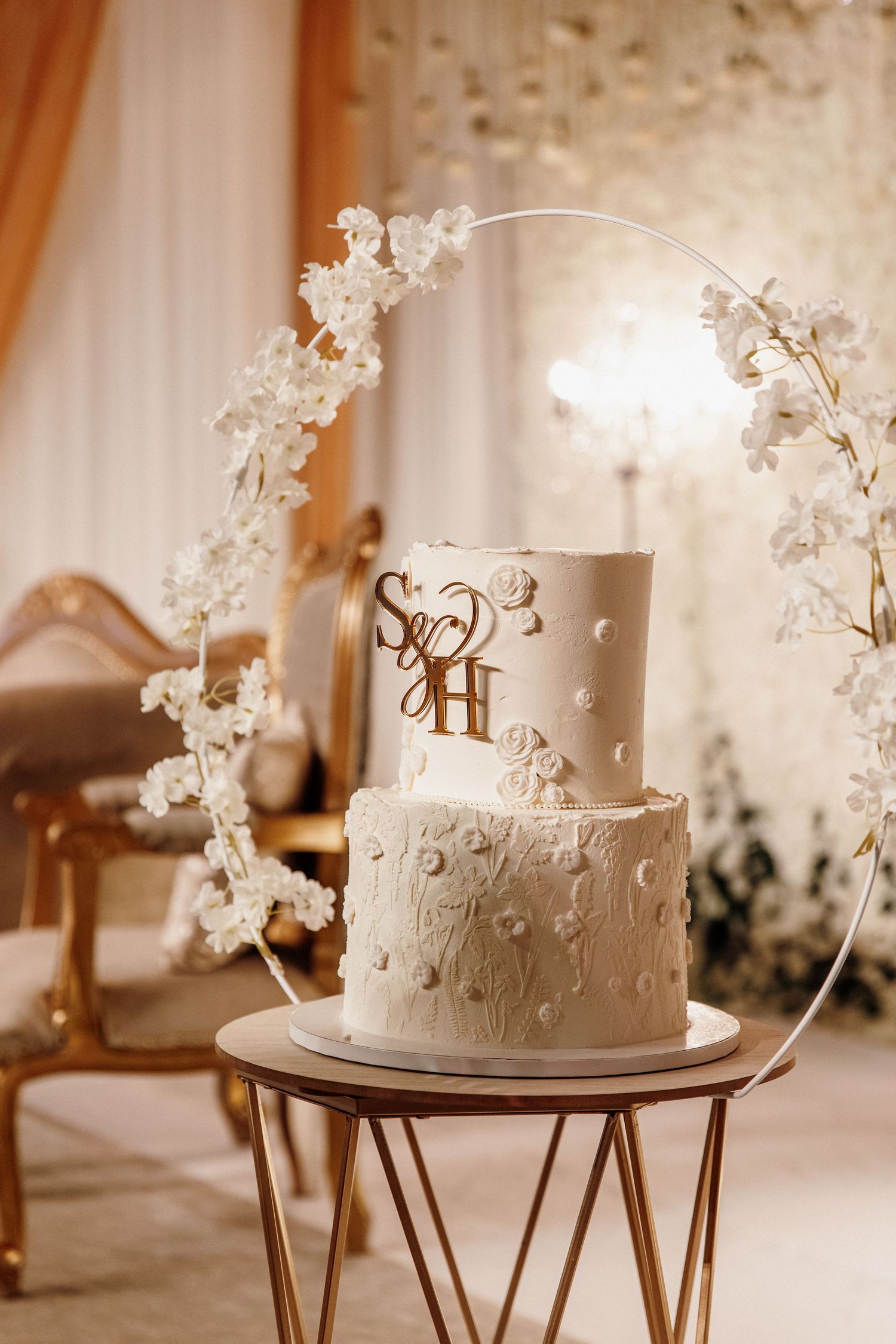 Elegant two-tier white wedding cake with floral details displayed on gold geometric stand with orchid arch decoration.
