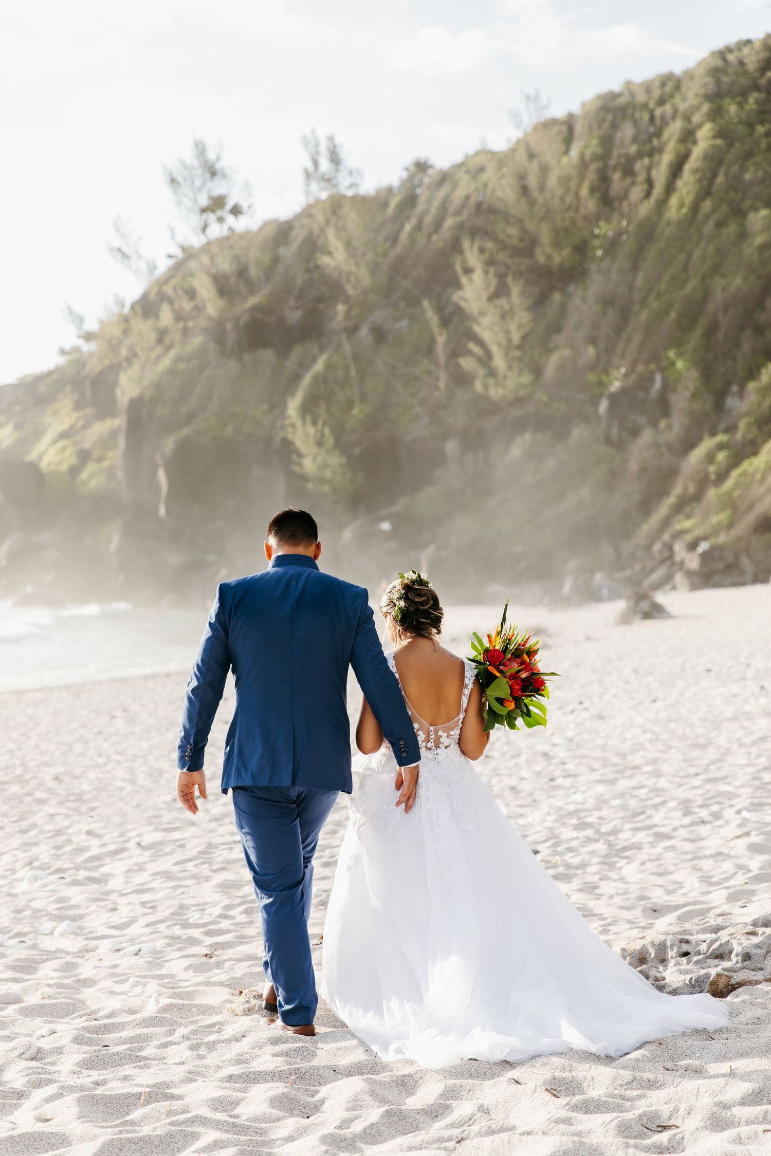 Newlyweds walking hand in hand on a scenic beach with dramatic cliffs in the background.