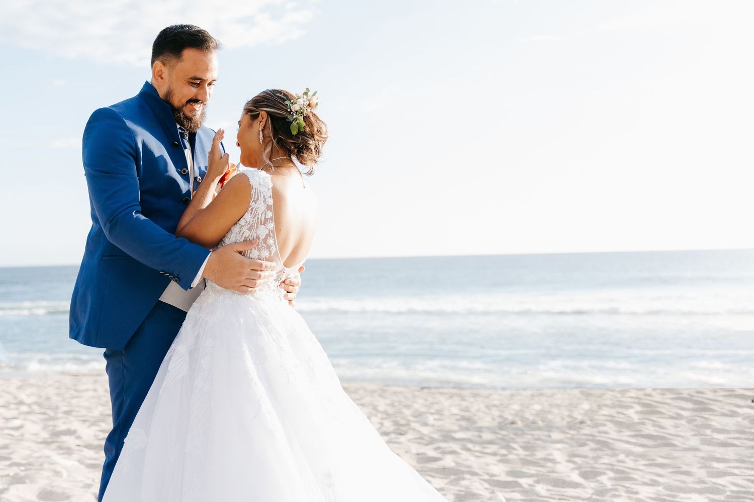 A romantic beach wedding moment captures a couple in white dress and blue suit against an ocean backdrop.