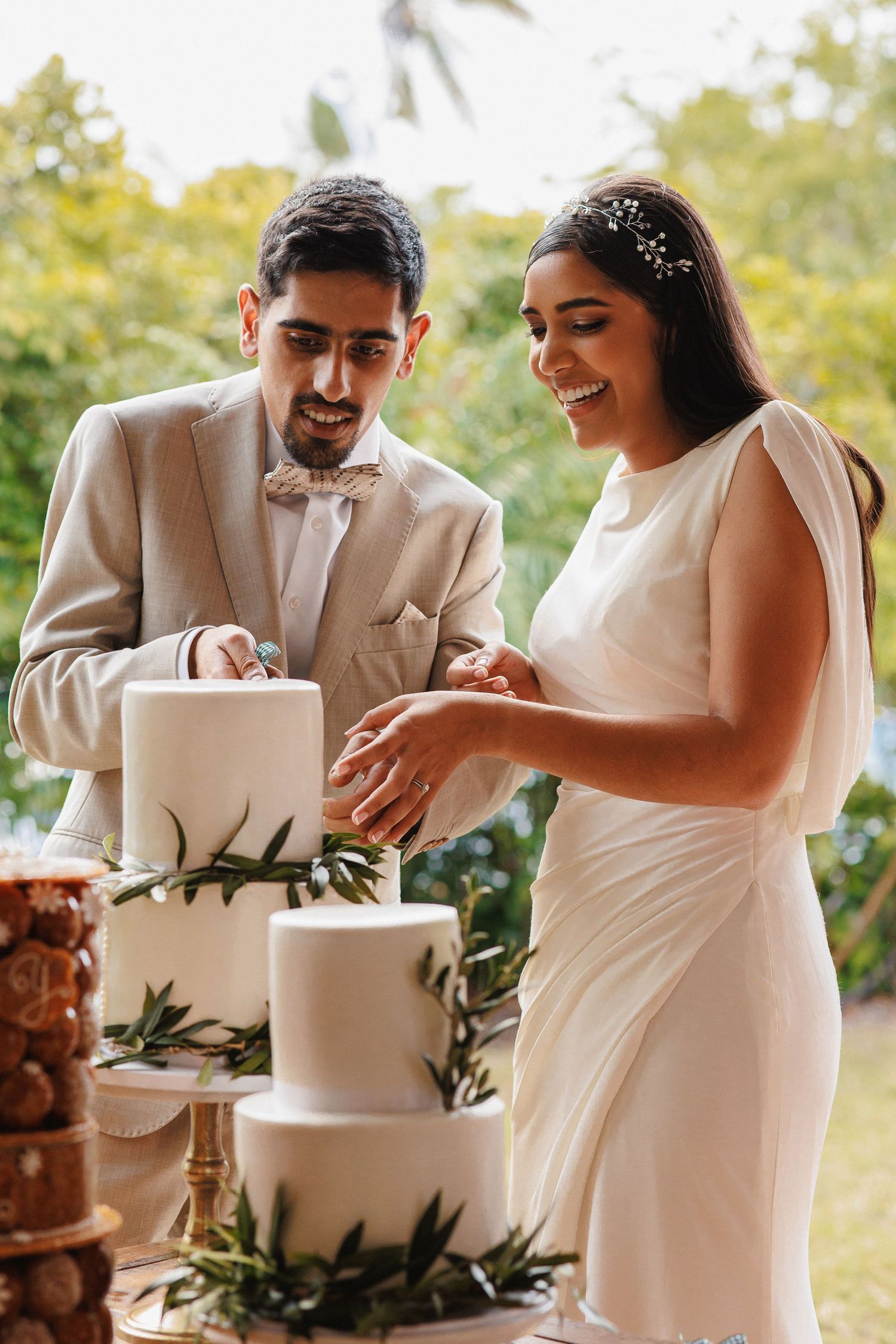 A bride and groom cutting their elegant white tiered wedding cake decorated with greenery.