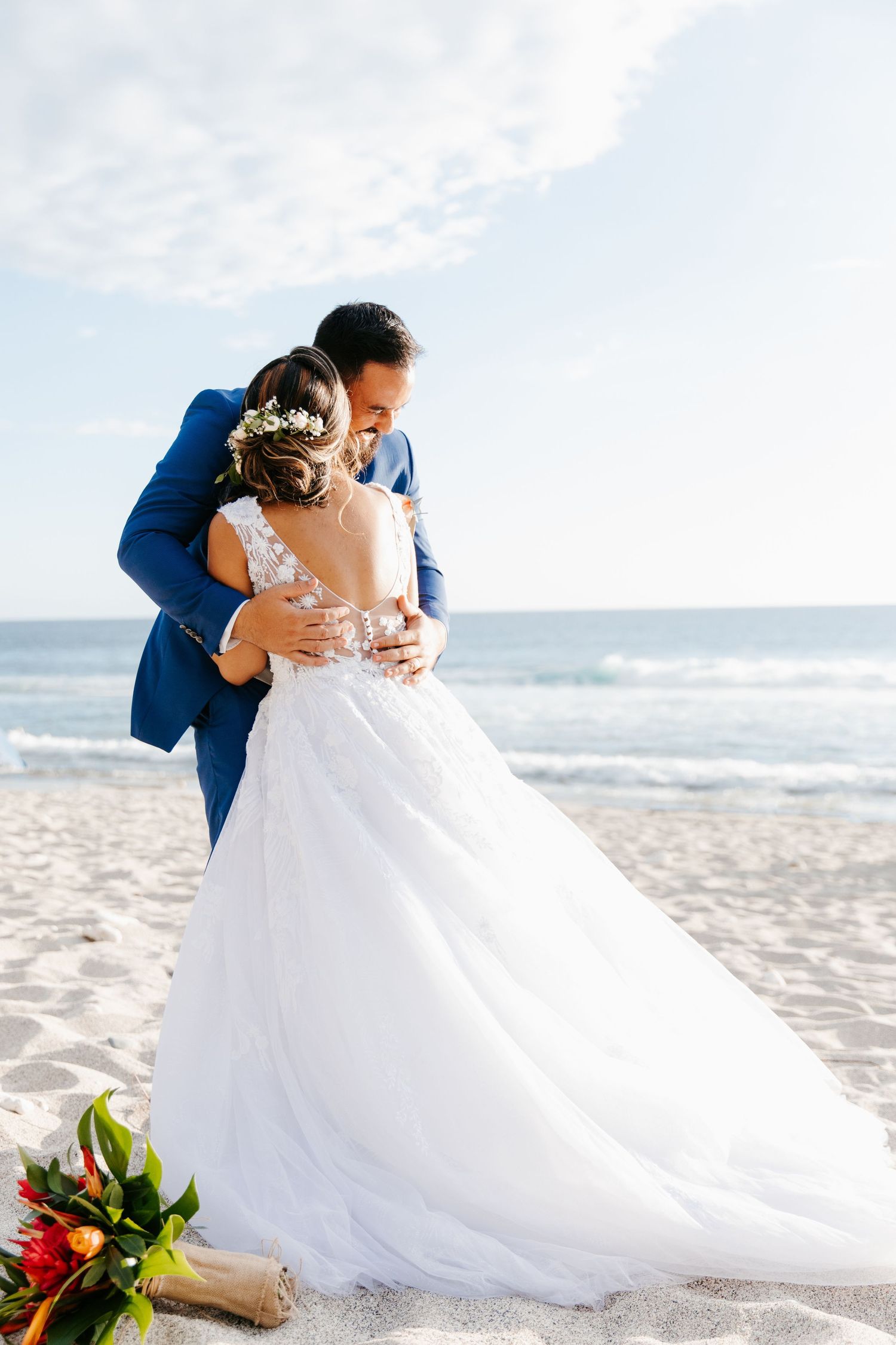 A romantic beach wedding photoshoot shows a couple embracing on white sand with a tropical bouquet at their feet.