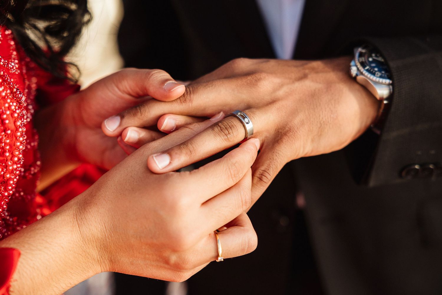 Close-up shot of hands exchanging wedding rings with detailed red sleeve in frame.