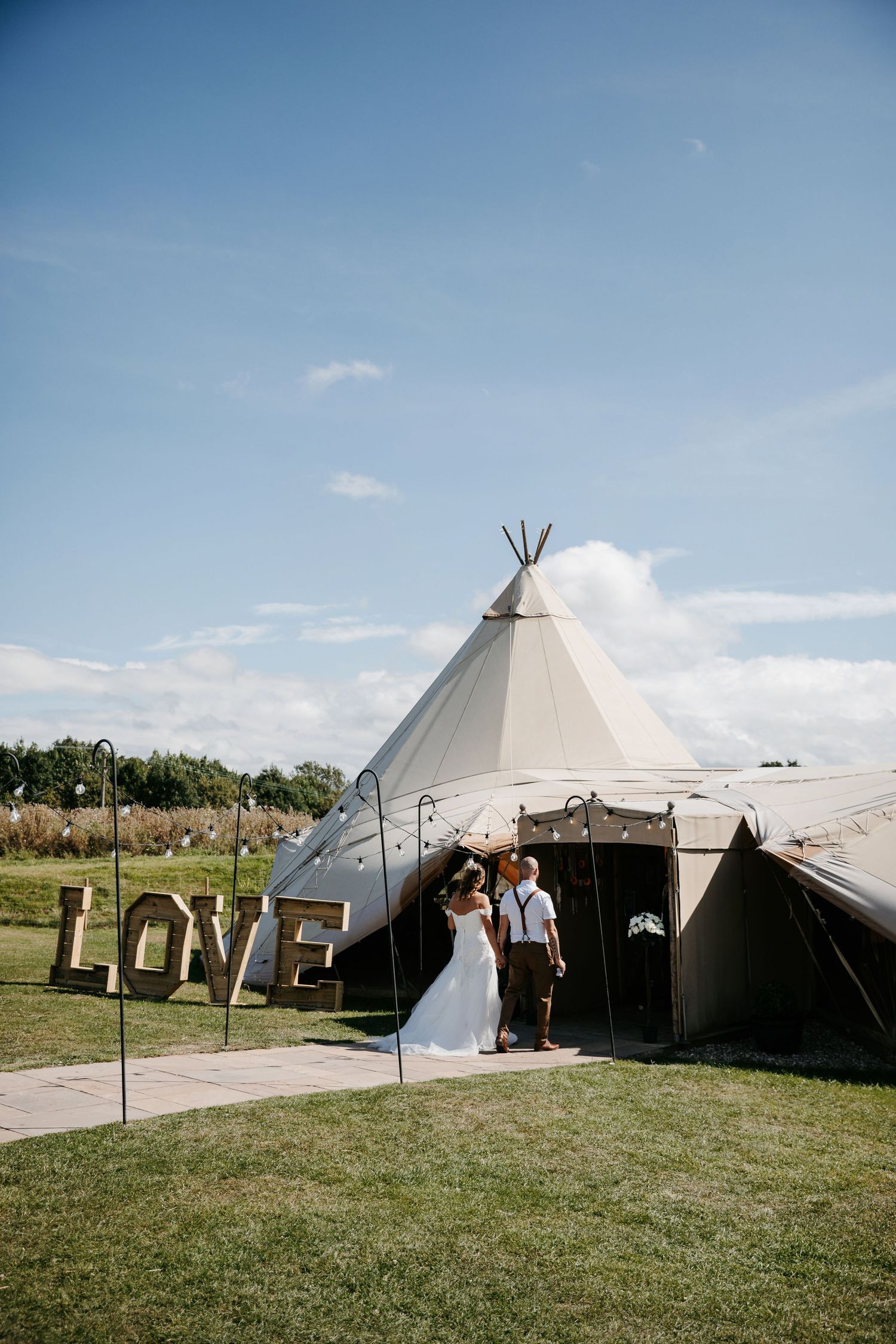 Wedding setup with large teepee tent and LOVE sign on grassy field under blue sky.