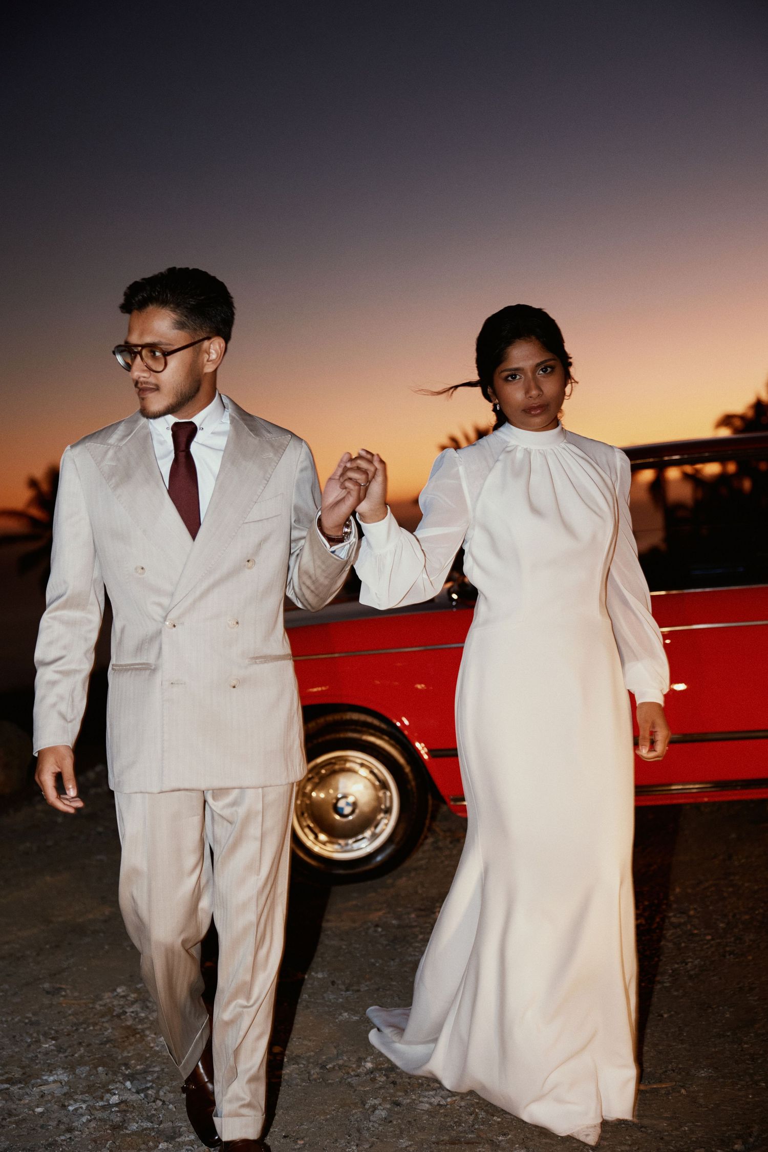 Elegant couple in white formal wear walking together at dusk beside a classic red car.