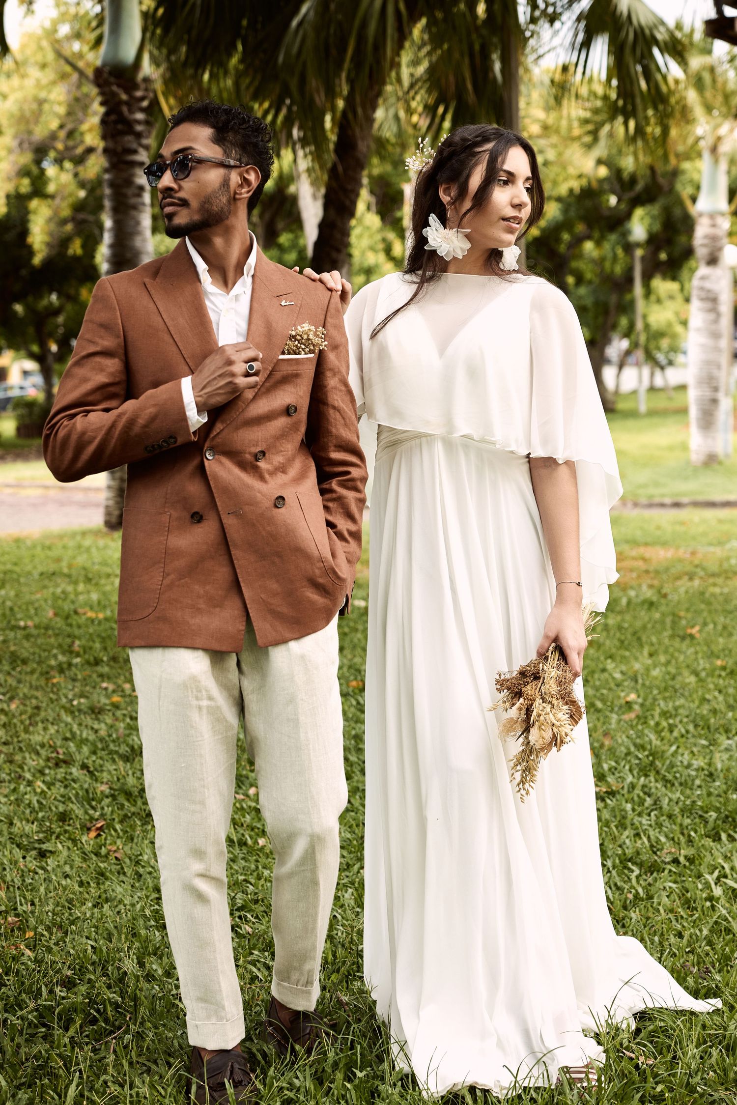A stylish couple in wedding attire pose in a palm tree garden setting.