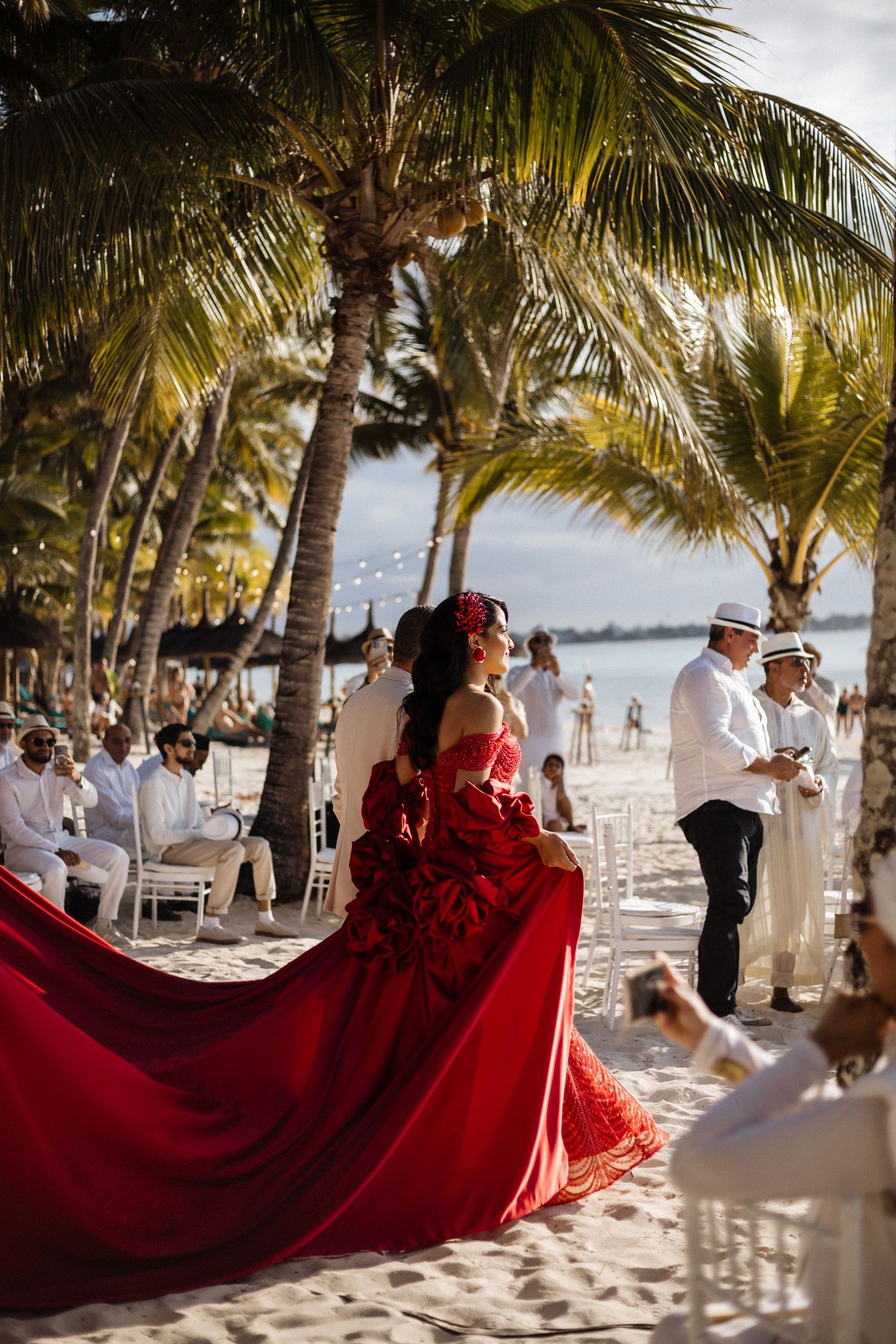 A dramatic red wedding dress with a long train flows across the sand at a beach ceremony.