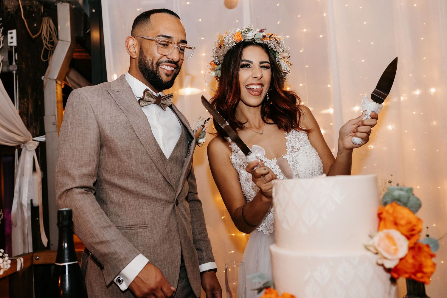 A joyful couple cutting their elegant white wedding cake decorated with orange roses at their reception.