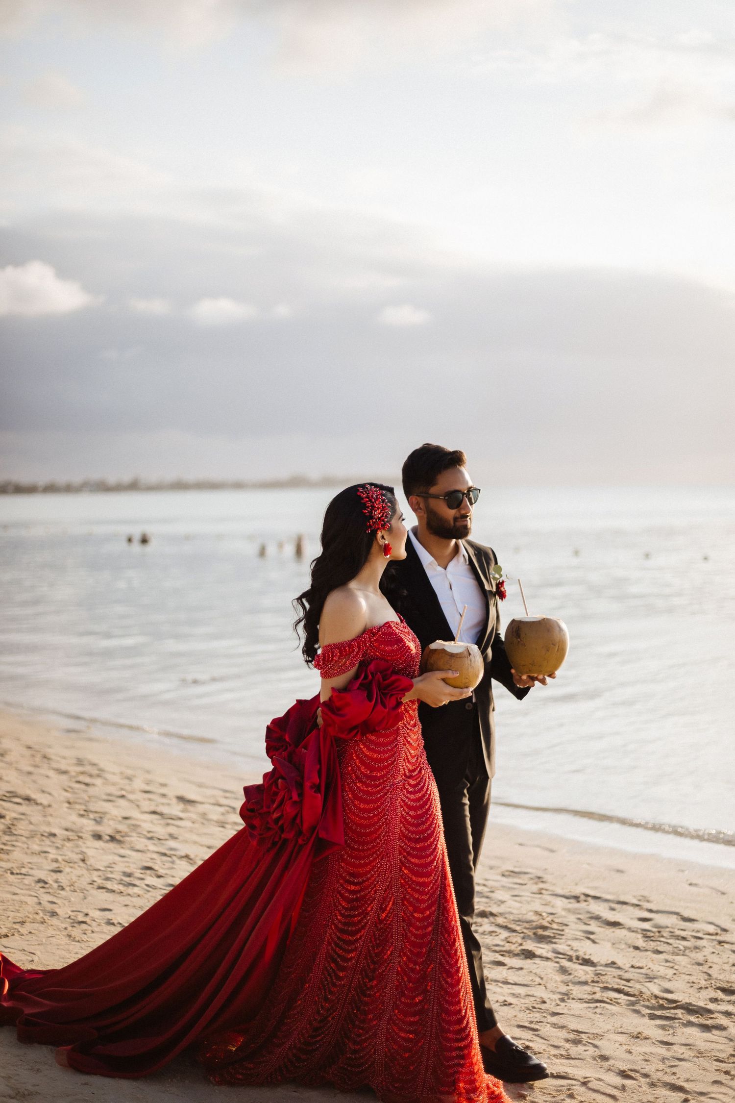 Couple enjoys coconut drinks on beach, bride in flowing red dress and groom in classic black suit.