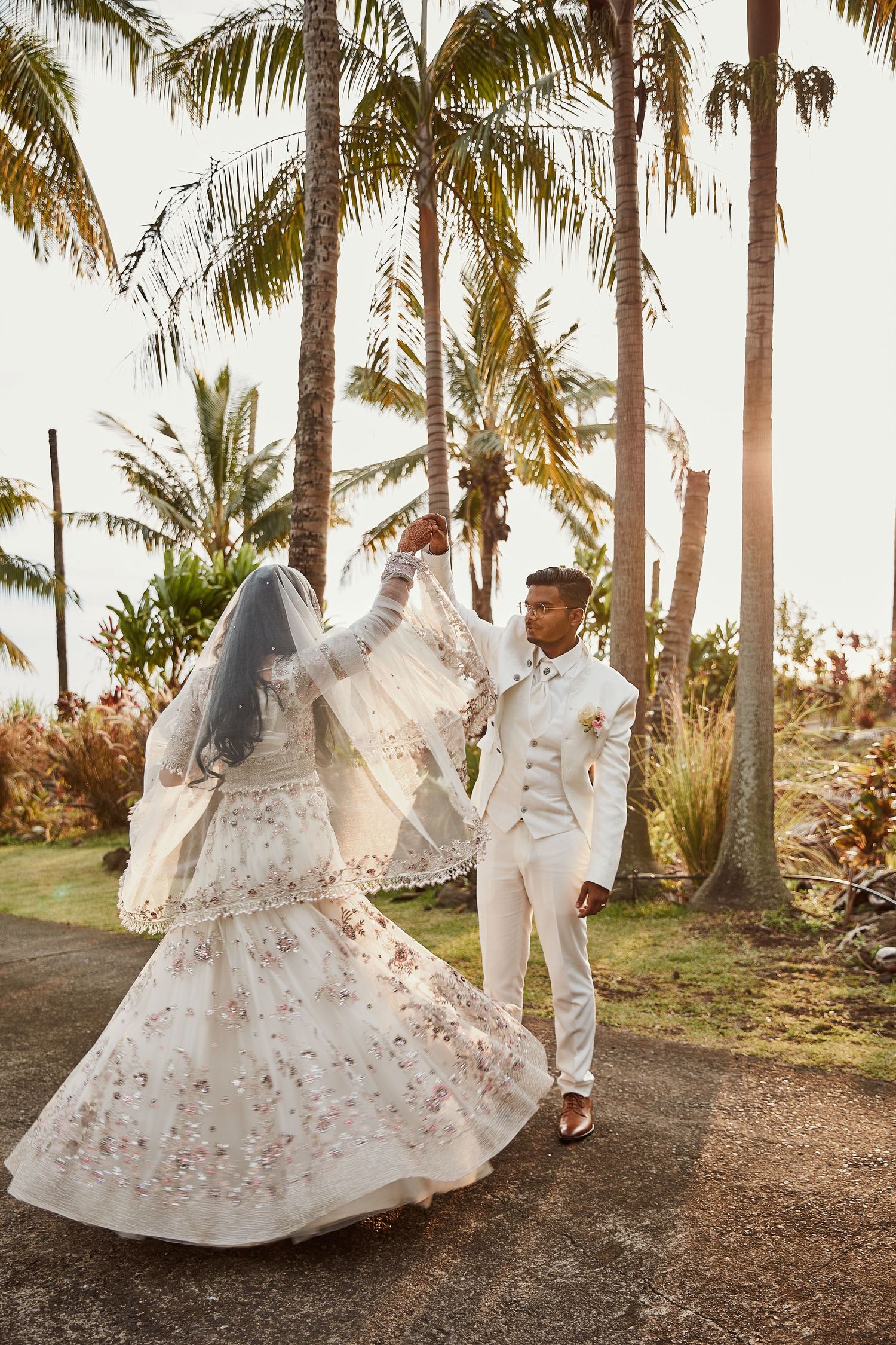 Couple dancing under palm trees during sunset in elegant white wedding attire.
