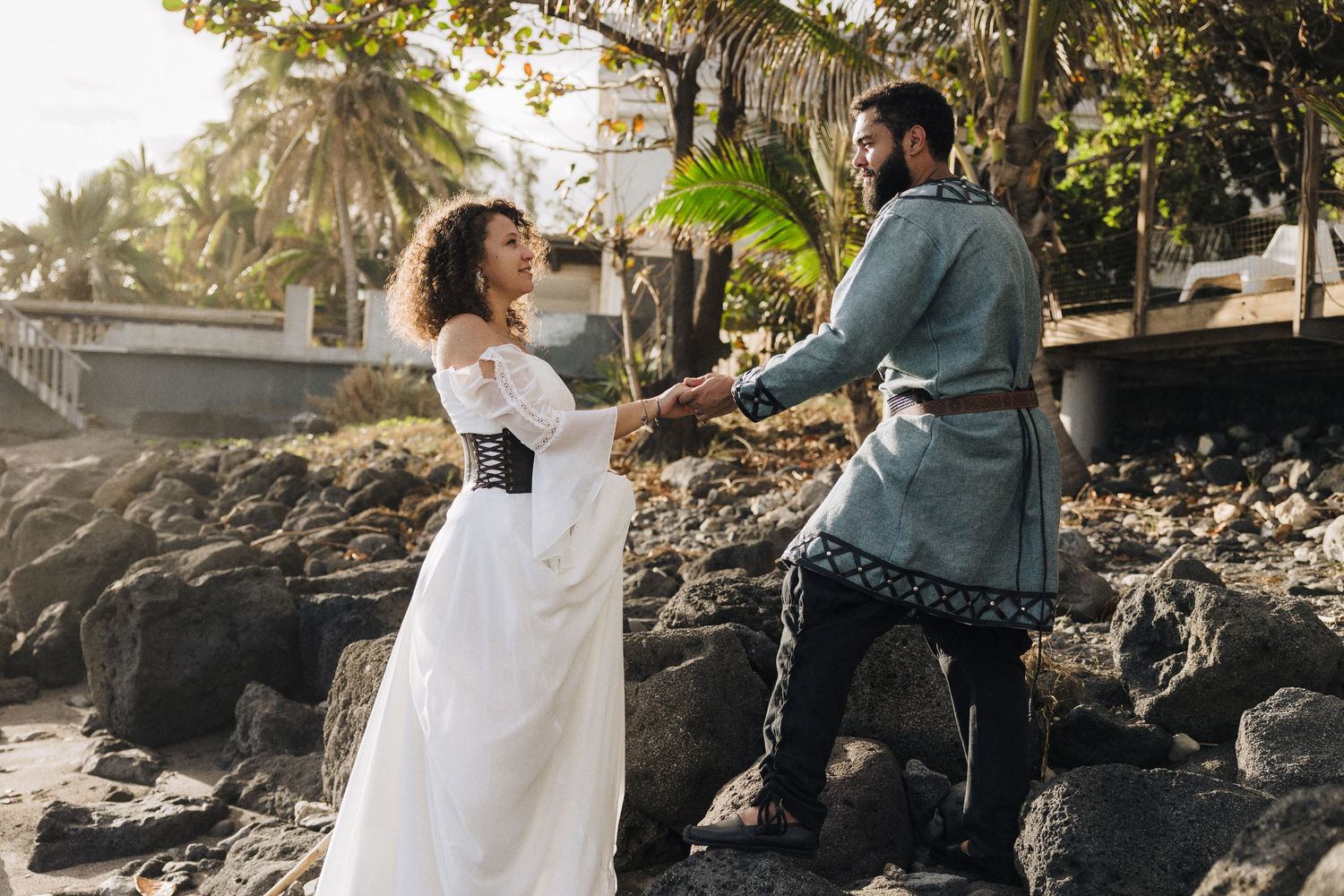 Couple in formal attire poses outdoors with palm trees and natural lighting in a romantic photoshoot setting.