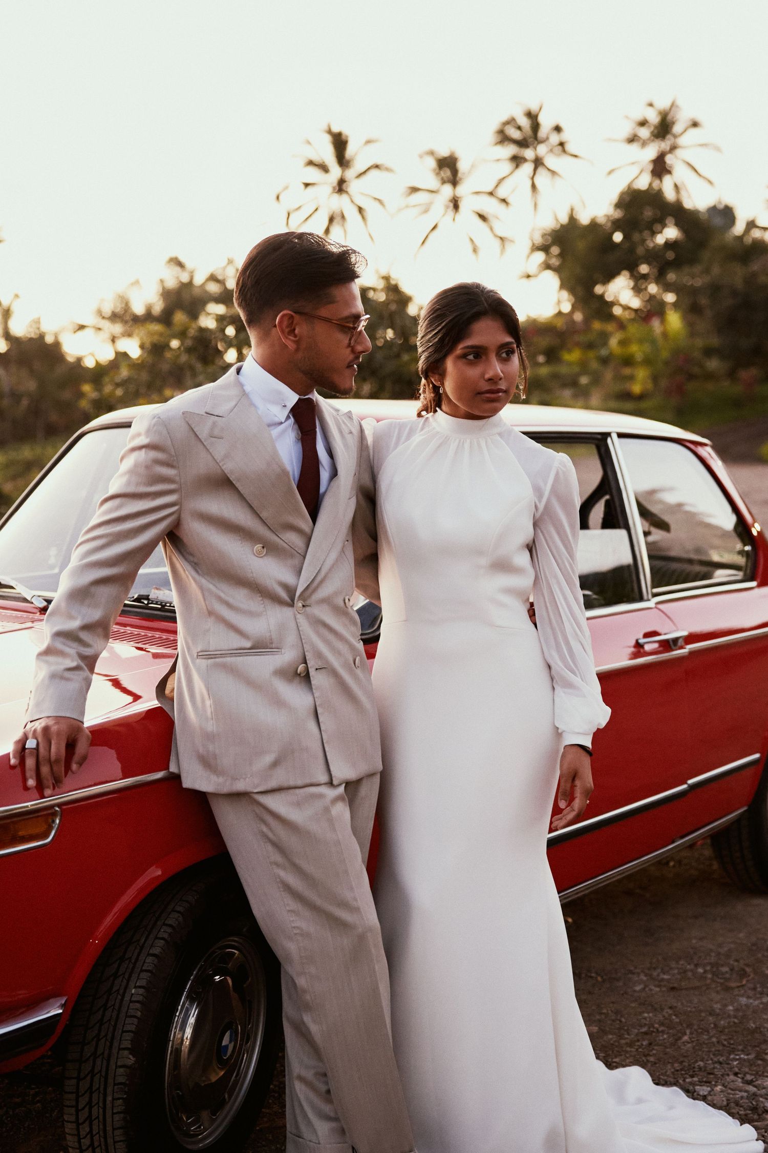 Stylish couple in white poses next to classic red car at sunset with palm trees in background.