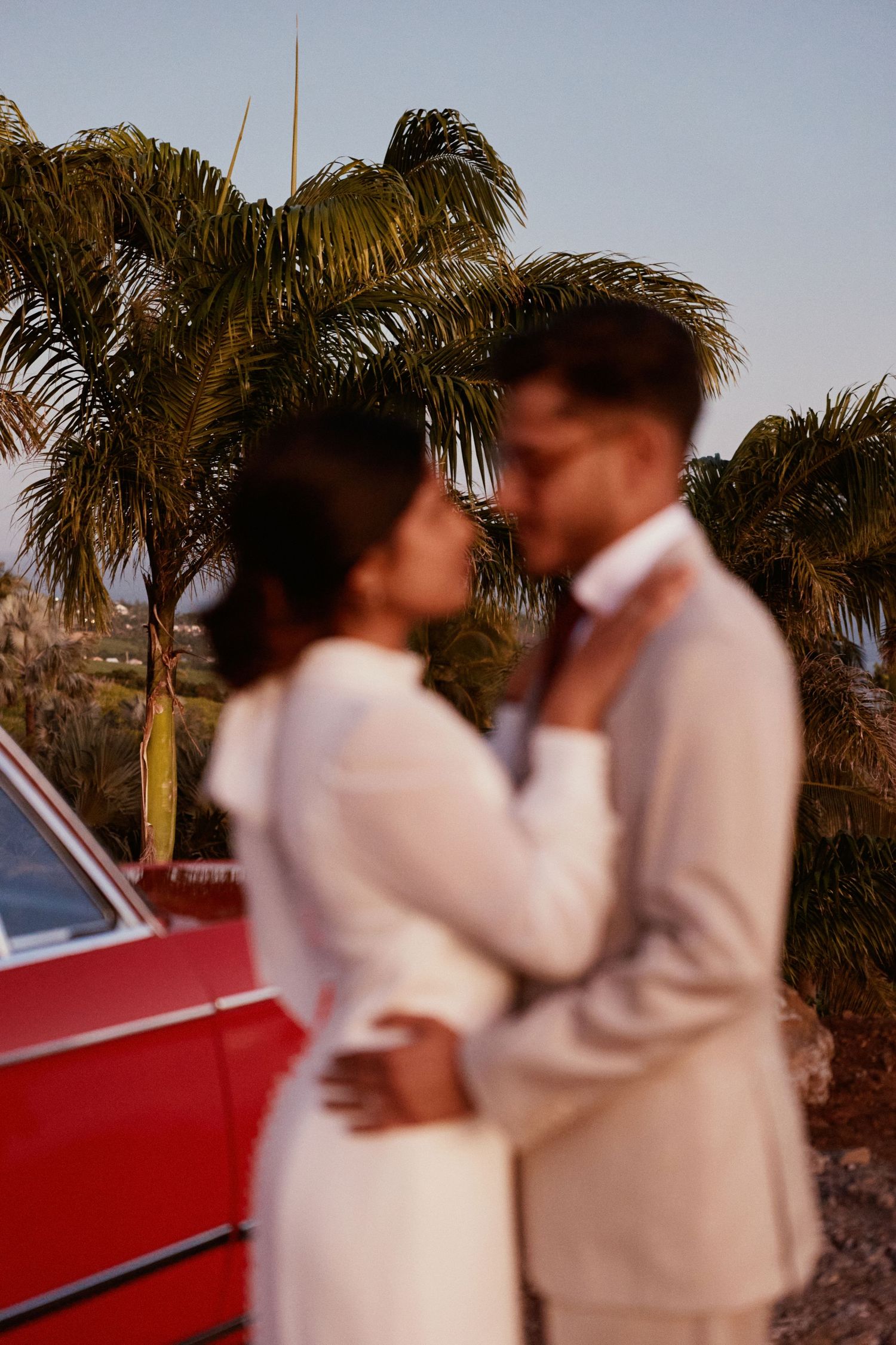 Romantic sunset portrait of a couple embracing near a vintage red car and palm trees.