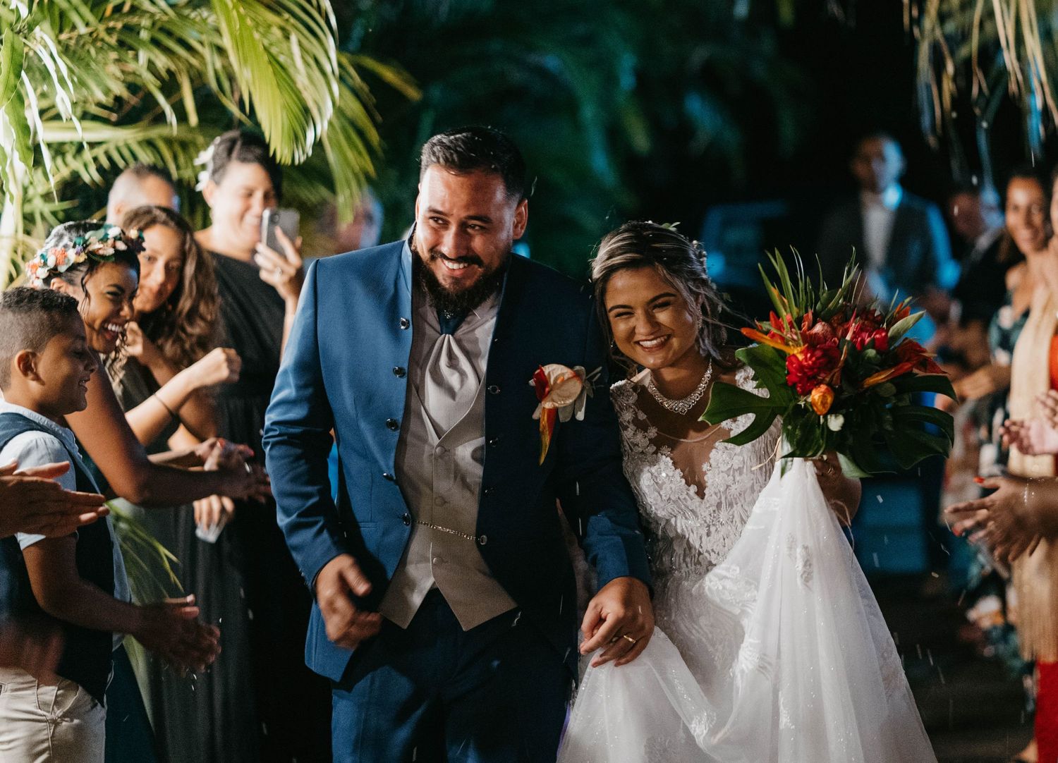 A wedding reception party scene with guests dancing under outdoor lighting and palm trees.
