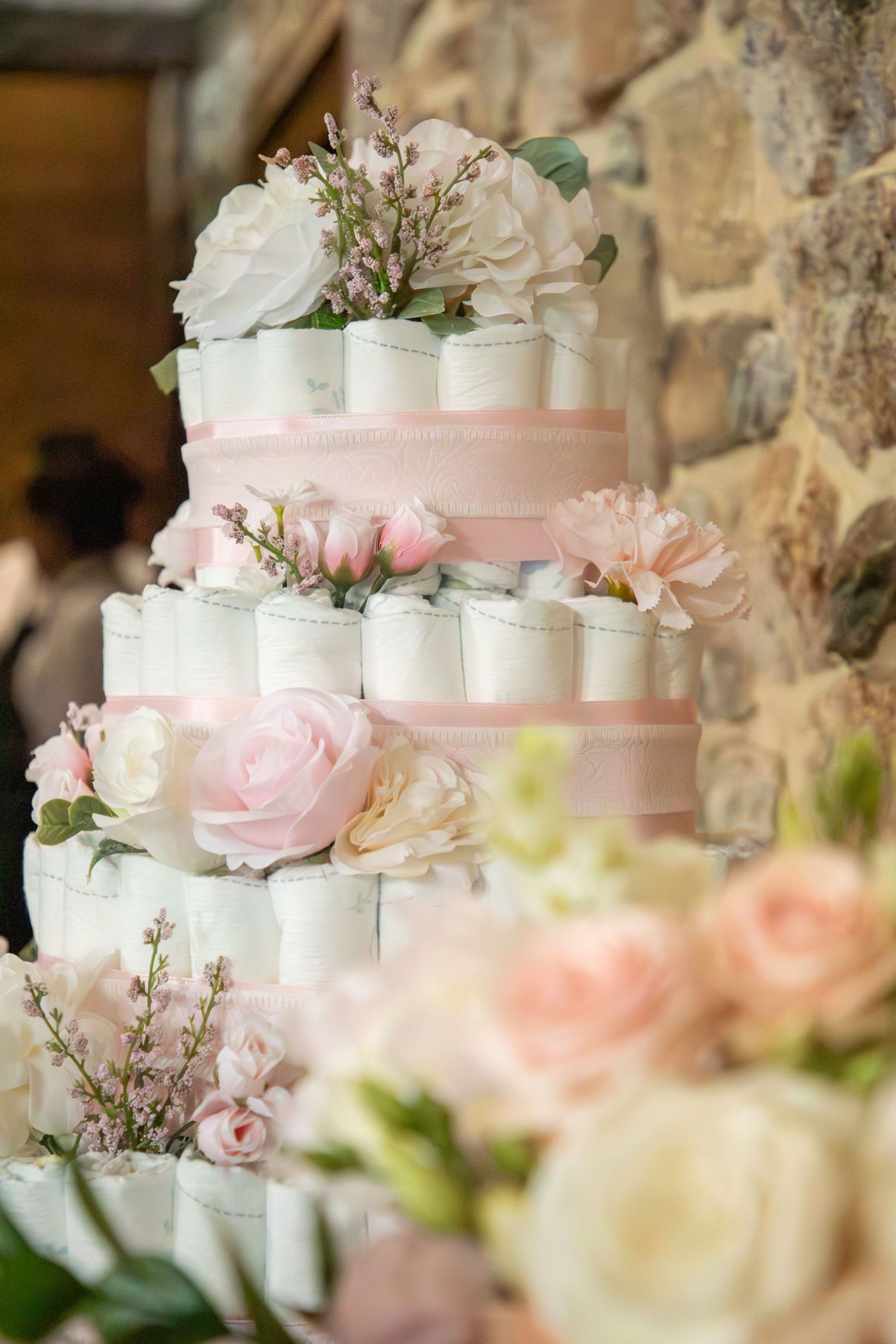 A tiered diaper cake decorated with blush pink ribbons and fresh roses in white and pink tones against a stone wall.