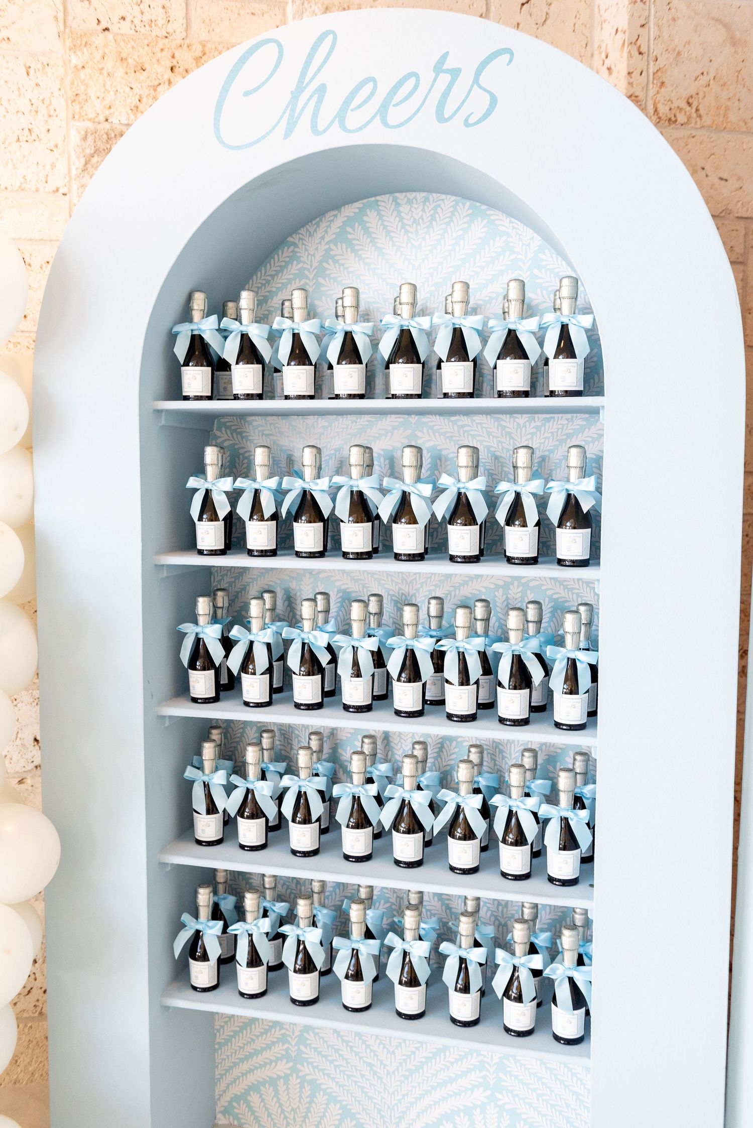 Mini champagne bottles arranged in rows on white arched display shelves with 'Cheers' text at the top.