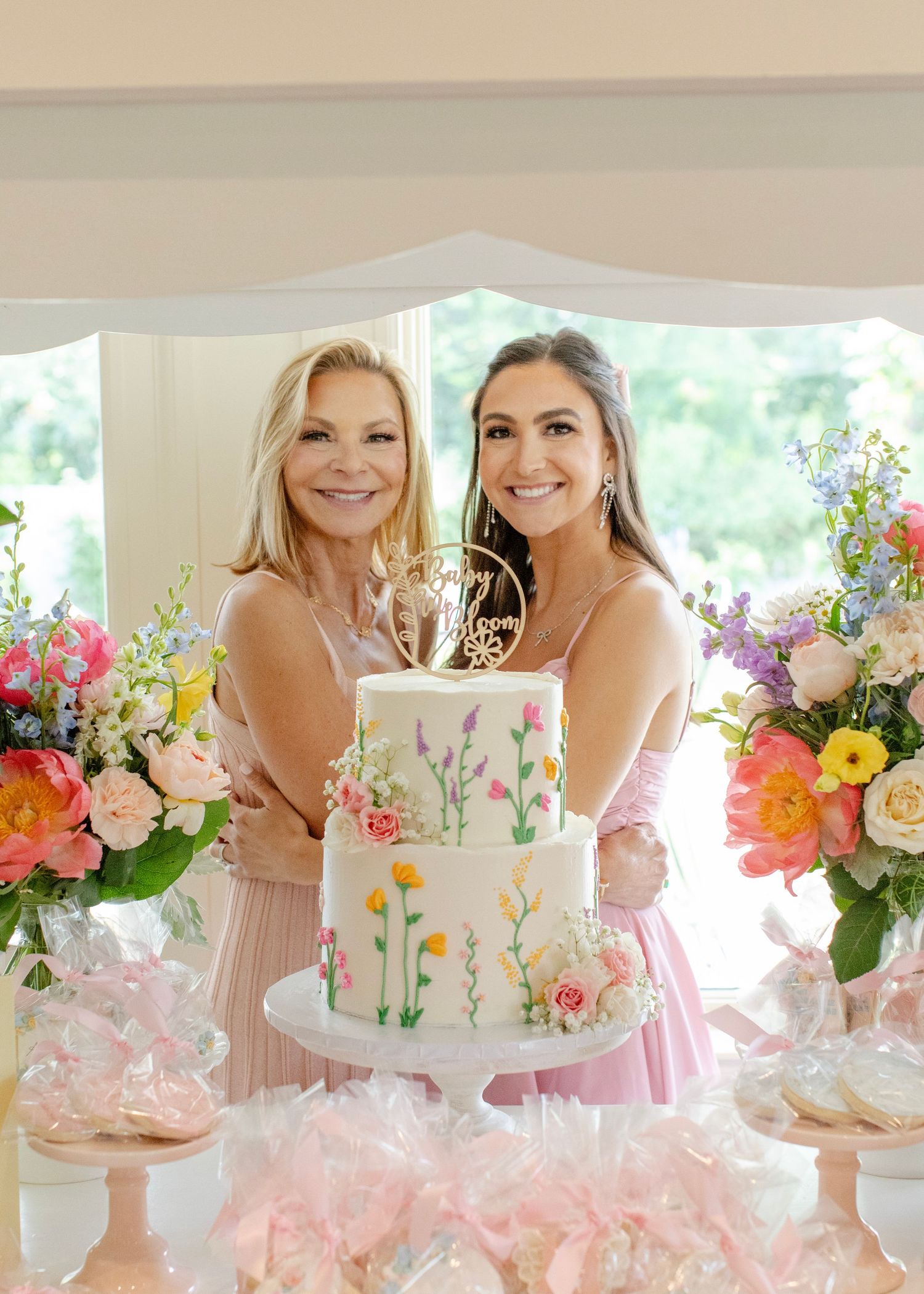 Mommy and daughter pose behind pink and blush floral party decorations with cakes and desserts.