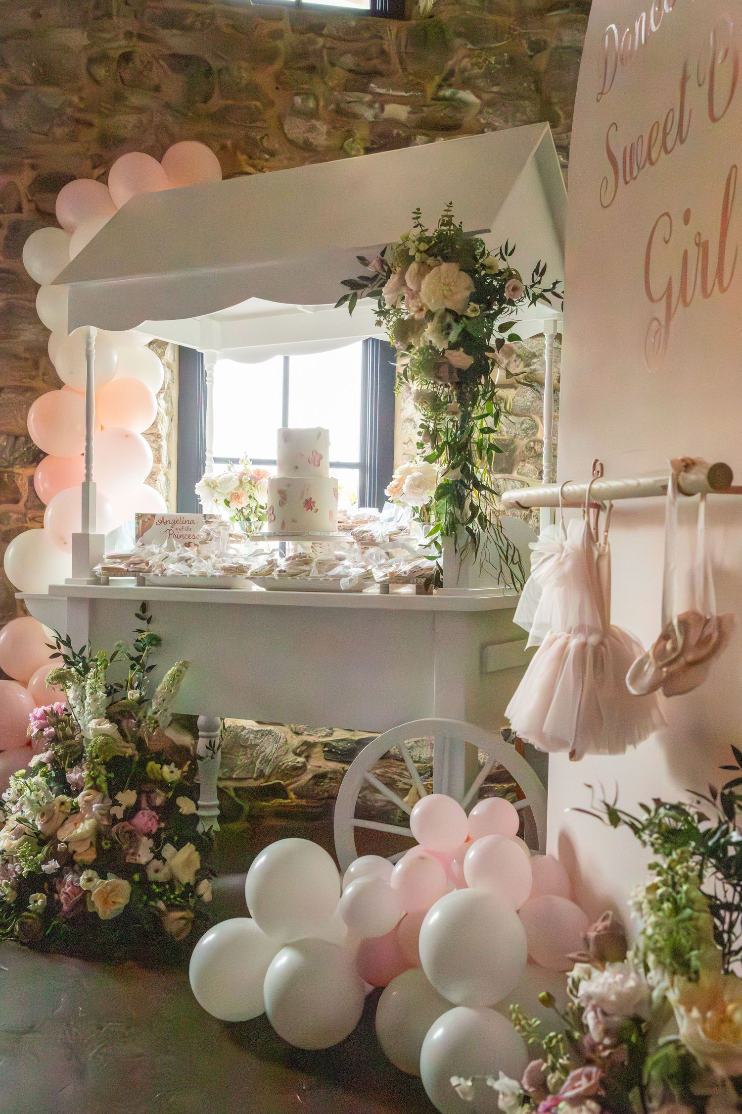 Party decor featuring a sweet cart, pink and white balloons, floral arrangements, and tulle dresses displayed in a rustic stone room.