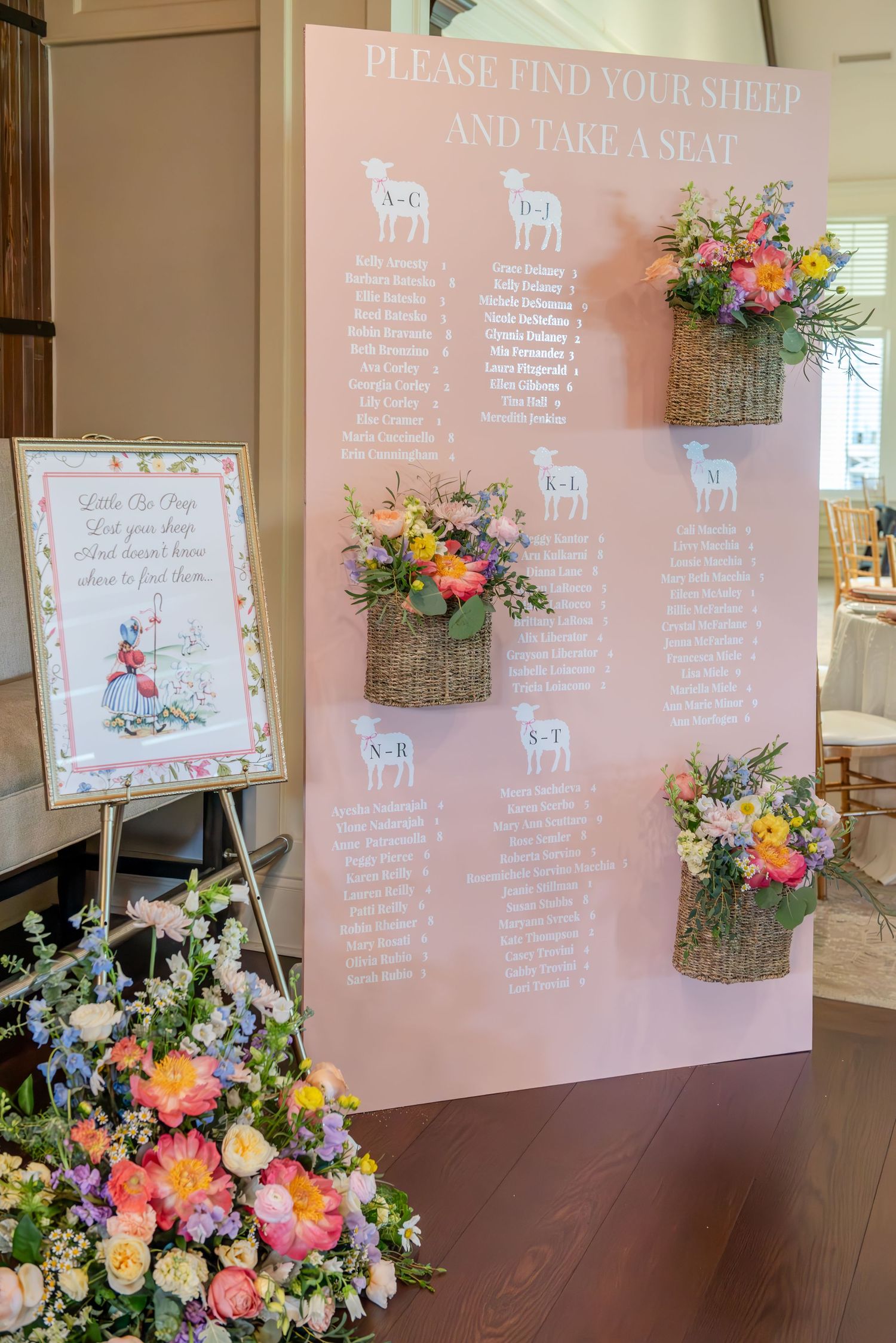 Bridal shower seating chart display with pink backdrop and floral arrangements in rustic baskets featuring colorful blooms.