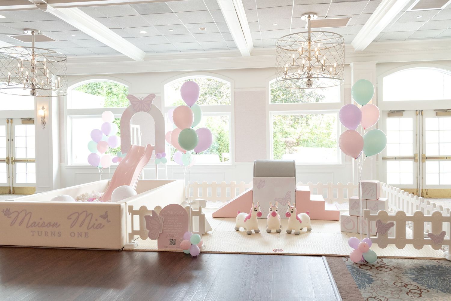 Pastel pink and mint party setup with balloons, decorative blocks, and elegant chandeliers in a bright white room.