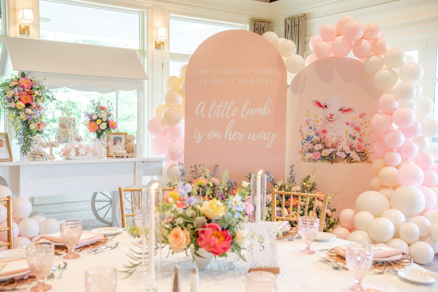A whimsical baby shower celebration setup with pink balloons, floral arrangements and elegant table settings in a bright room.