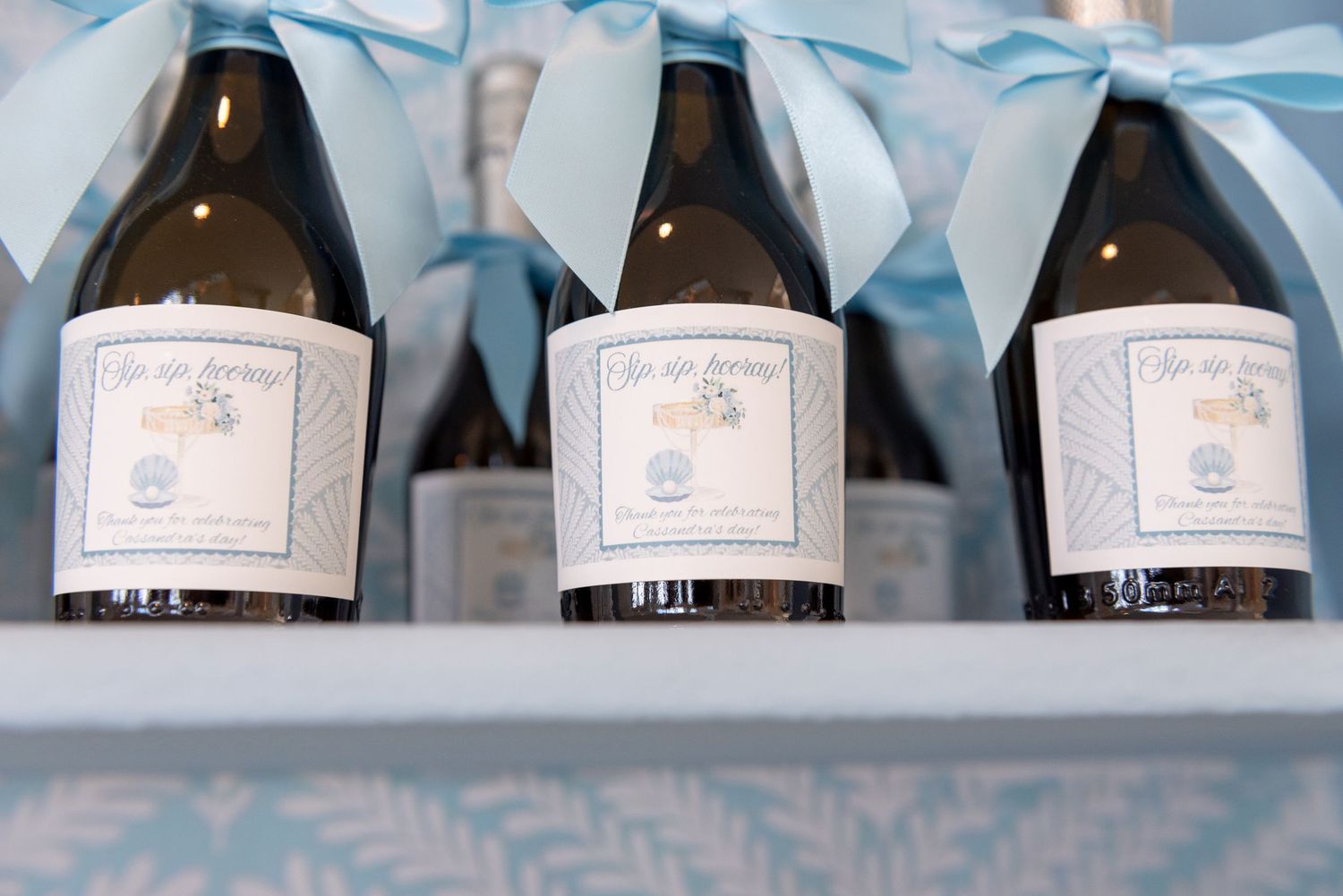 Three champagne bottles with light blue ribbons and custom labels displayed in a row on a white surface.