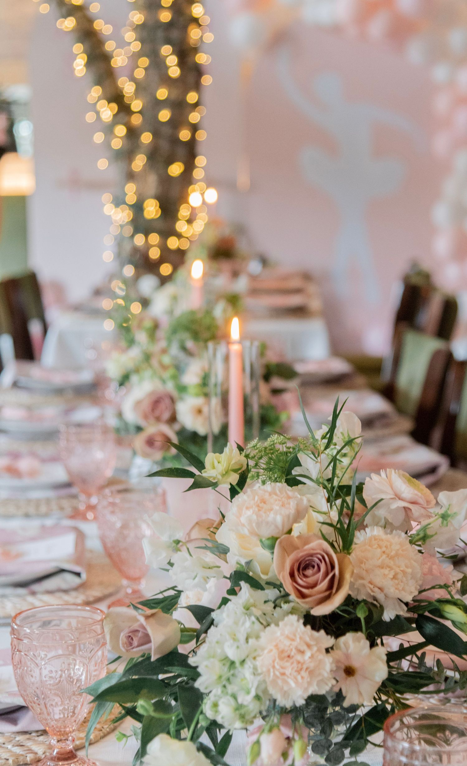Elegant baby shower table settings with pink and white floral centerpieces and glowing string lights in the background.